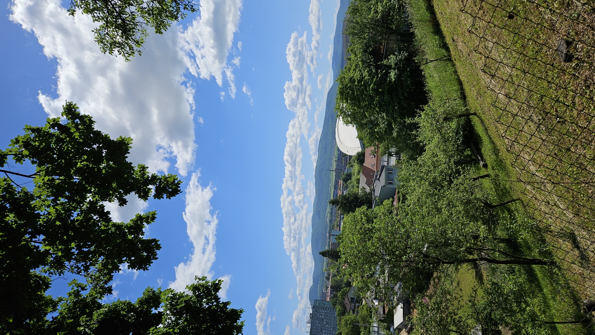 The image presents a view from an elevated position in Prešov, Slovakia, overlooking a landscape encompassing both urban and natural elements. In the foreground, foliage from trees frames the scene on the left and right sides. The central portion of the view showcases a vibrant blue sky with puffy white clouds, stretching towards a distant mountain range.
Below the mountains, a cluster of buildings, including residential houses and taller apartment blocks, forms part of the cityscape. A large, white satellite dish is visible among the buildings, suggesting the presence of a telecommunications or scientific facility. On the right side, a chain-link fence lines a grassy slope, creating a visual boundary between the elevated viewpoint and the lower-lying areas.