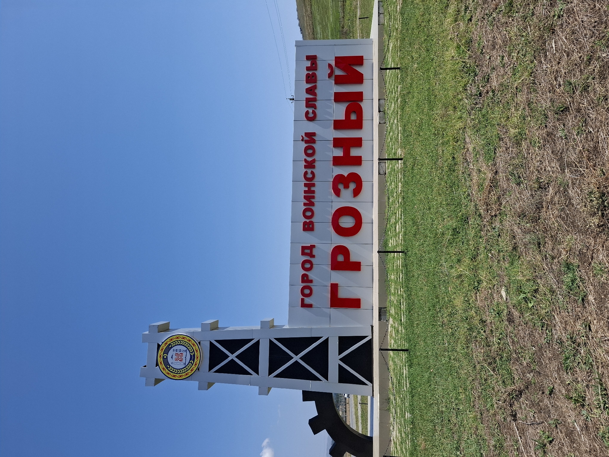 The image shows a large, modern monument with prominent red lettering against a clear blue sky. The monument features a tall, white, tower-like structure on the left, adorned with a circular emblem at the top and crisscrossing black patterns lower down. To the right of the tower is a large white wall with the words "ГОРОД ВОИНСКОЙ СЛАВЫ" (City of Military Glory) in red lettering on the top line, and "ГРОЗНЫЙ" (Grozny) in larger red lettering on the bottom line. A large, black, gear-like structure is partially visible to the far left of the monument. The monument is situated on a paved area with a checkered pattern of grey stones, bordered by a low black chain fence. In front of the paved area is a strip of green grass, which transitions into drier, sparser vegetation further down. The background shows a gentle, green hillside under a bright, cloudless sky. The location identified is the city of Pobedinskoye, Russia, which is known for being a gateway to Grozny.