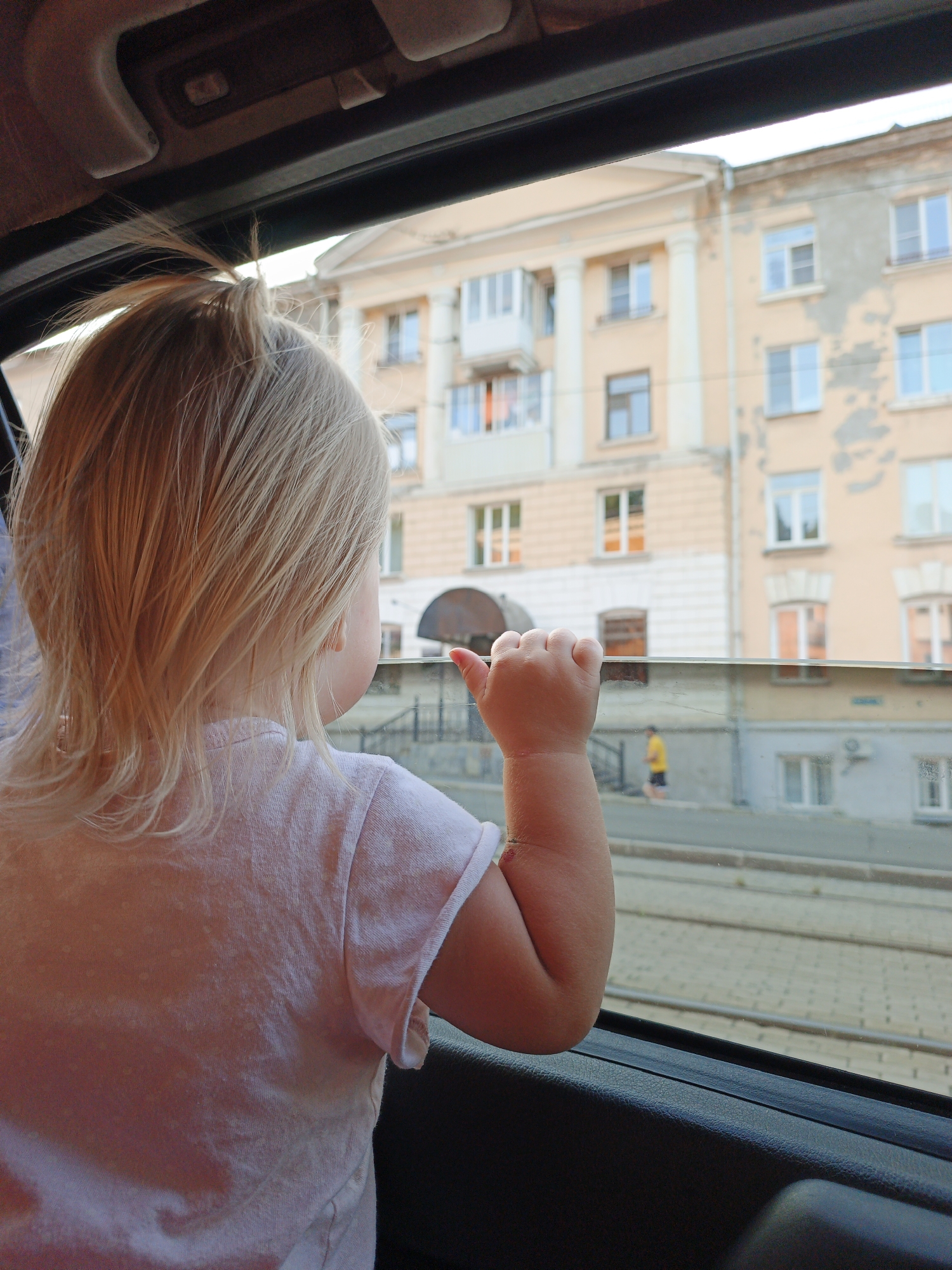 The image shows a young child with blond hair looking out of a car window. The child is wearing a pink t-shirt. Outside the window, there is a building with several windows and a balcony. In the background, a person in a yellow shirt is walking. The scene appears to be in a city, with cobblestone pavement visible in the foreground. The image is taken from inside a vehicle.