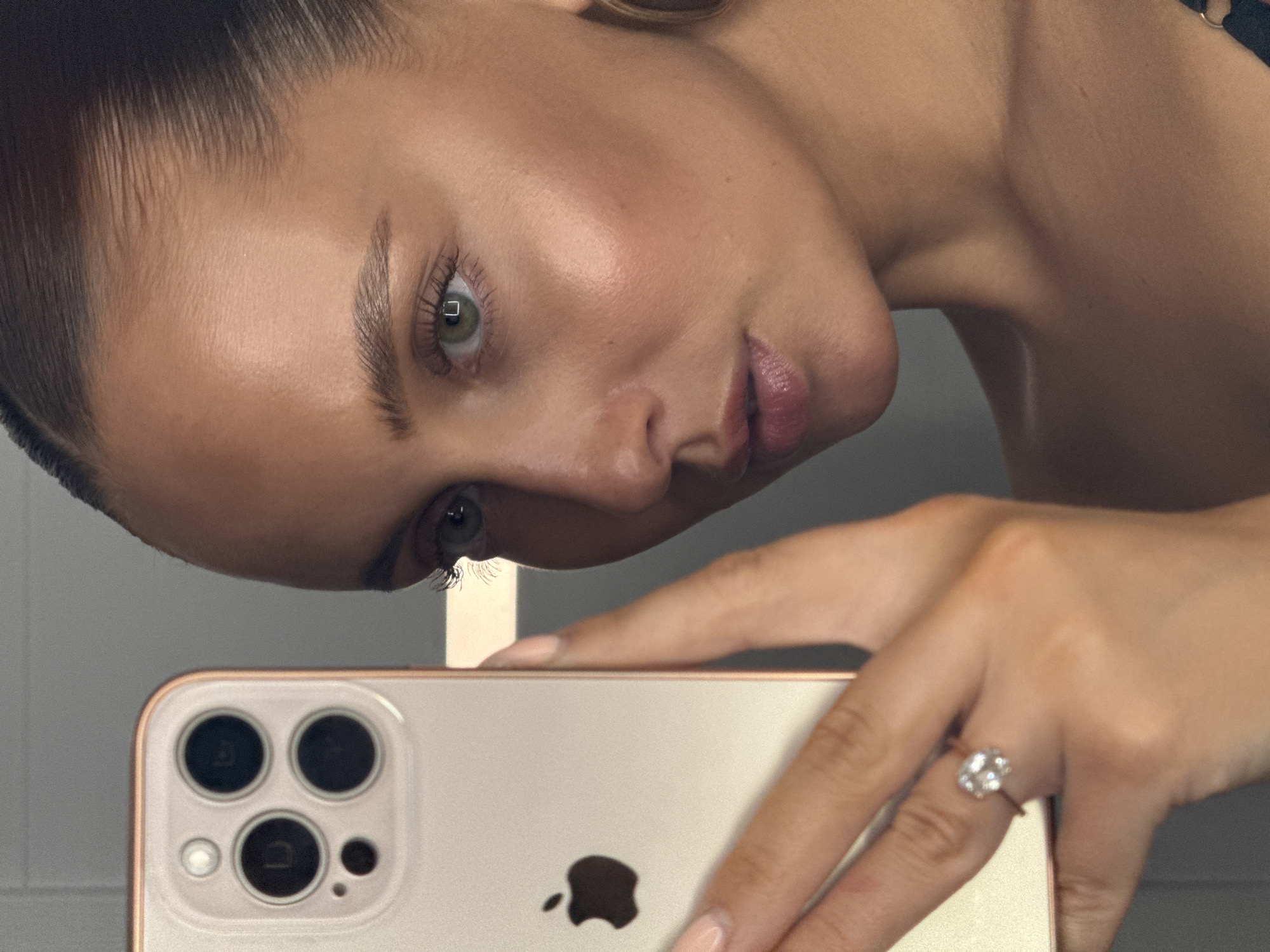 A woman is taking a selfie with her iPhone 13 Pro in the city of Chaikovsky, Russia. The woman is looking into the camera with her green eyes. Her hair is pulled back in a bun. She is wearing makeup and has a diamond ring on her left ring finger. The iPhone has a gold-colored case and is held in her left hand. The background is a gray wall.