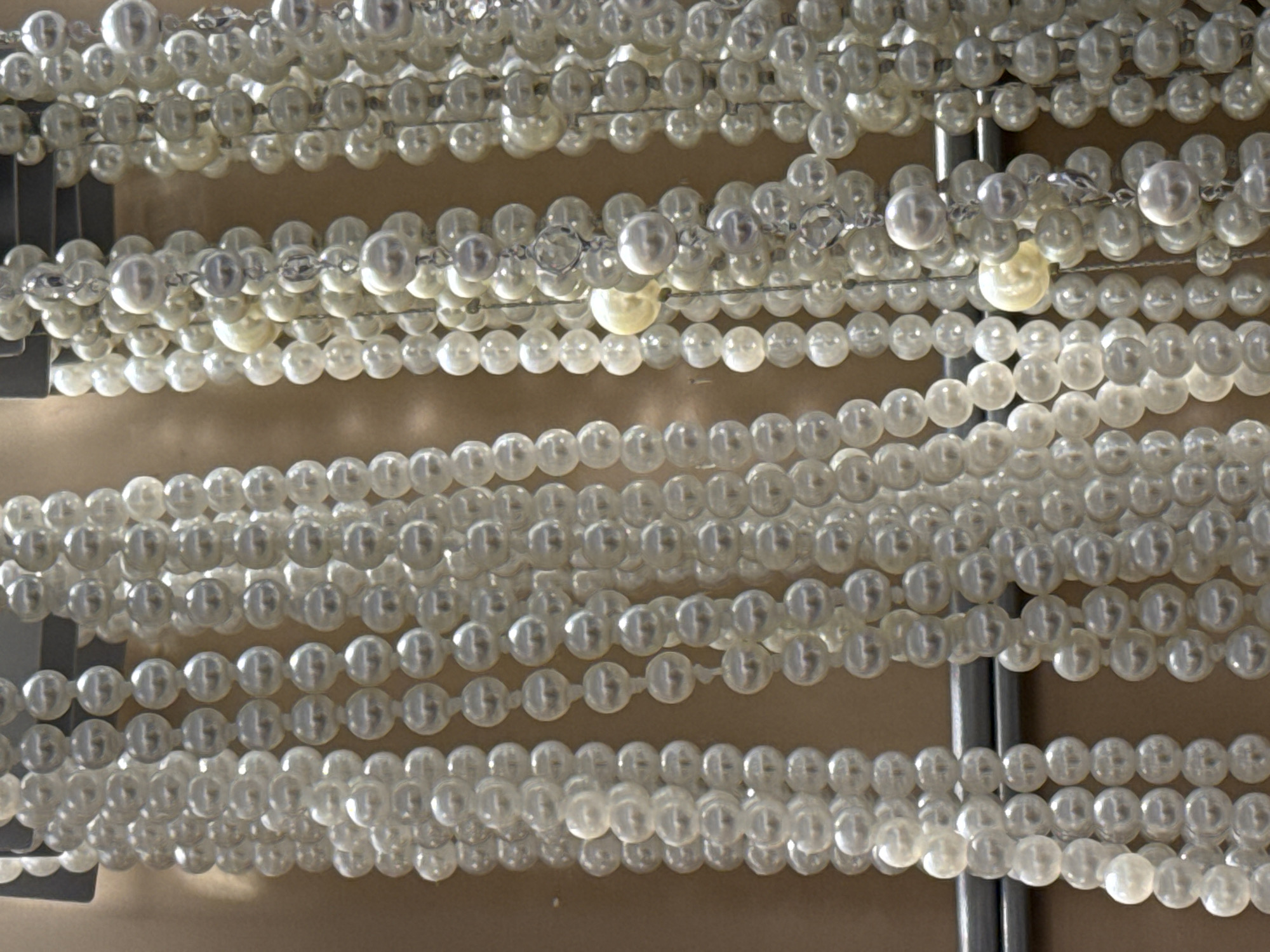 The image shows multiple strands of white pearl beads suspended from a gray metal frame. The beads are arranged in parallel lines, with some strands hanging lower than others. The background appears to be a light brown or tan color. The location is Bjeliši, Montenegro, but there is no location specific information in the image. There are no people present.