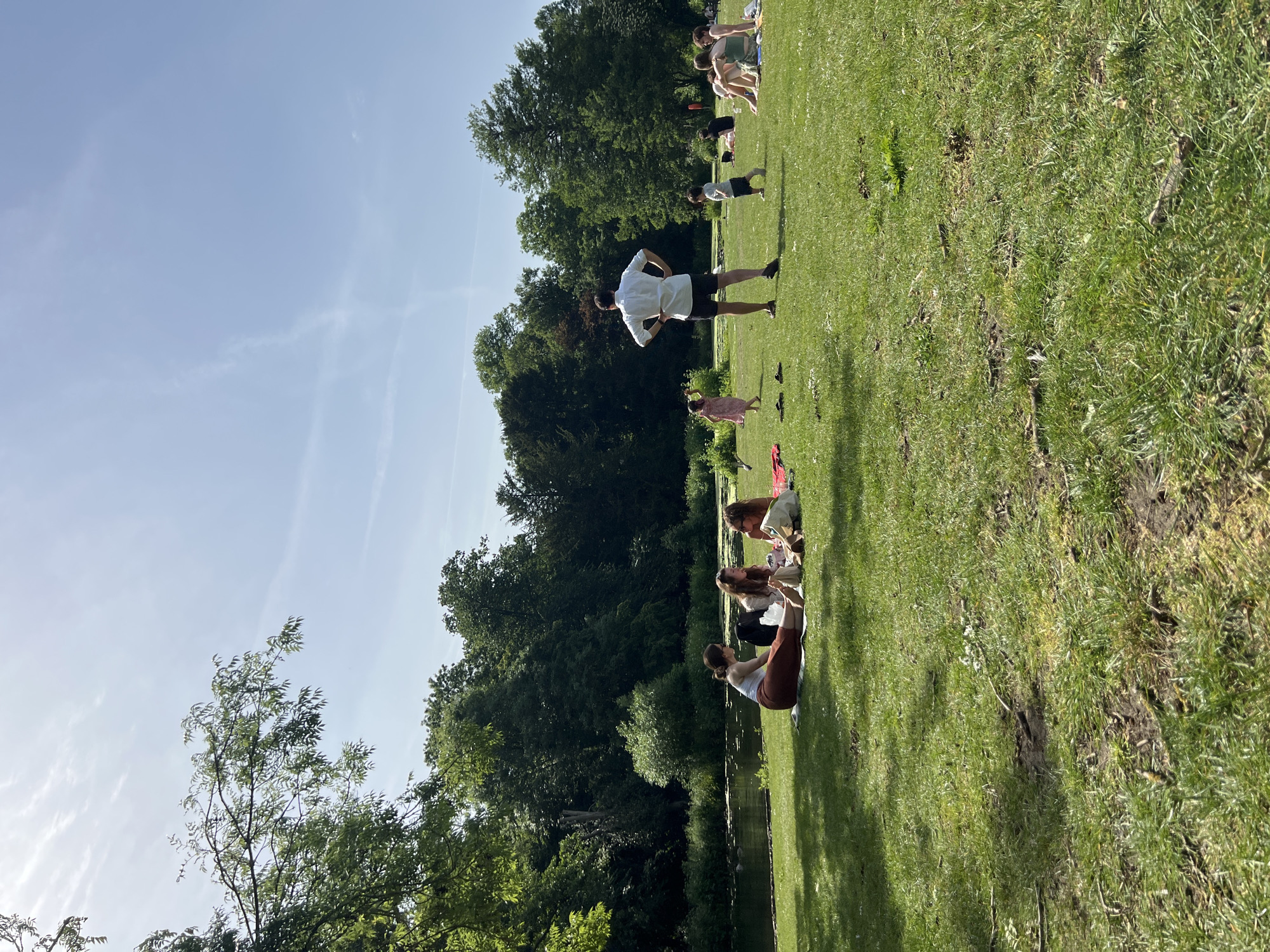 A group of people is gathered on a grassy area next to a body of water, likely a pond or lake. Some are seated, appearing to be in conversation or observing the scene. At least one individual is standing and seemingly instructing or observing the group. There are trees and greenery in the background, creating a park-like setting. The sky is visible in the upper portion of the image, suggesting it is daytime. There is a clear blue sky. The setting may be a park in London.