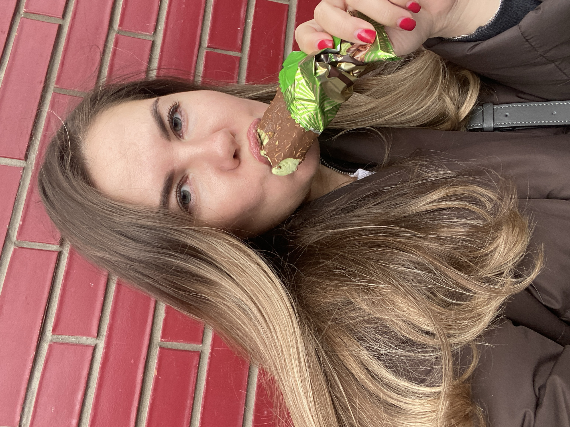 A fair-skinned woman with long, blonde hair and red nail polish is eating a chocolate-covered ice cream bar in front of a red brick wall. She is wearing a brown jacket. The location appears to be an urban area, potentially a sidewalk, with the red brick wall suggesting it could be the side of a building. Given the instructions, the location is presumed to be in Izhevsk, Russia.