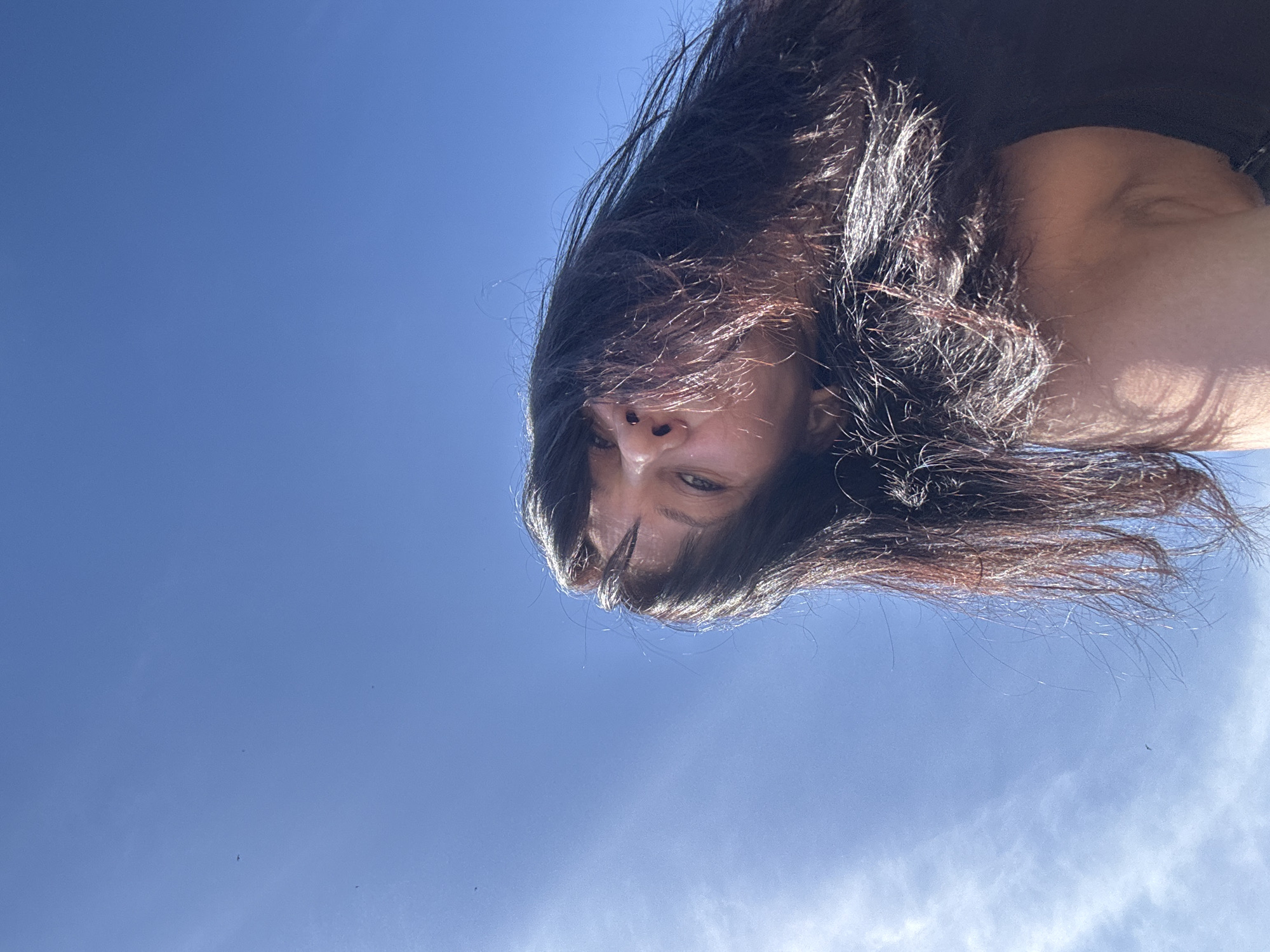 A low-angle shot captures a person with long, dark hair against a bright blue sky. The person's face is visible, with a focus on their eyes and nose. The hair is disheveled, with strands blowing in various directions. The background is a clear blue sky with some wisps of cloud in the lower right corner. There are no discernible landmarks to determine the location within the city of Chaikovsky, Russia.