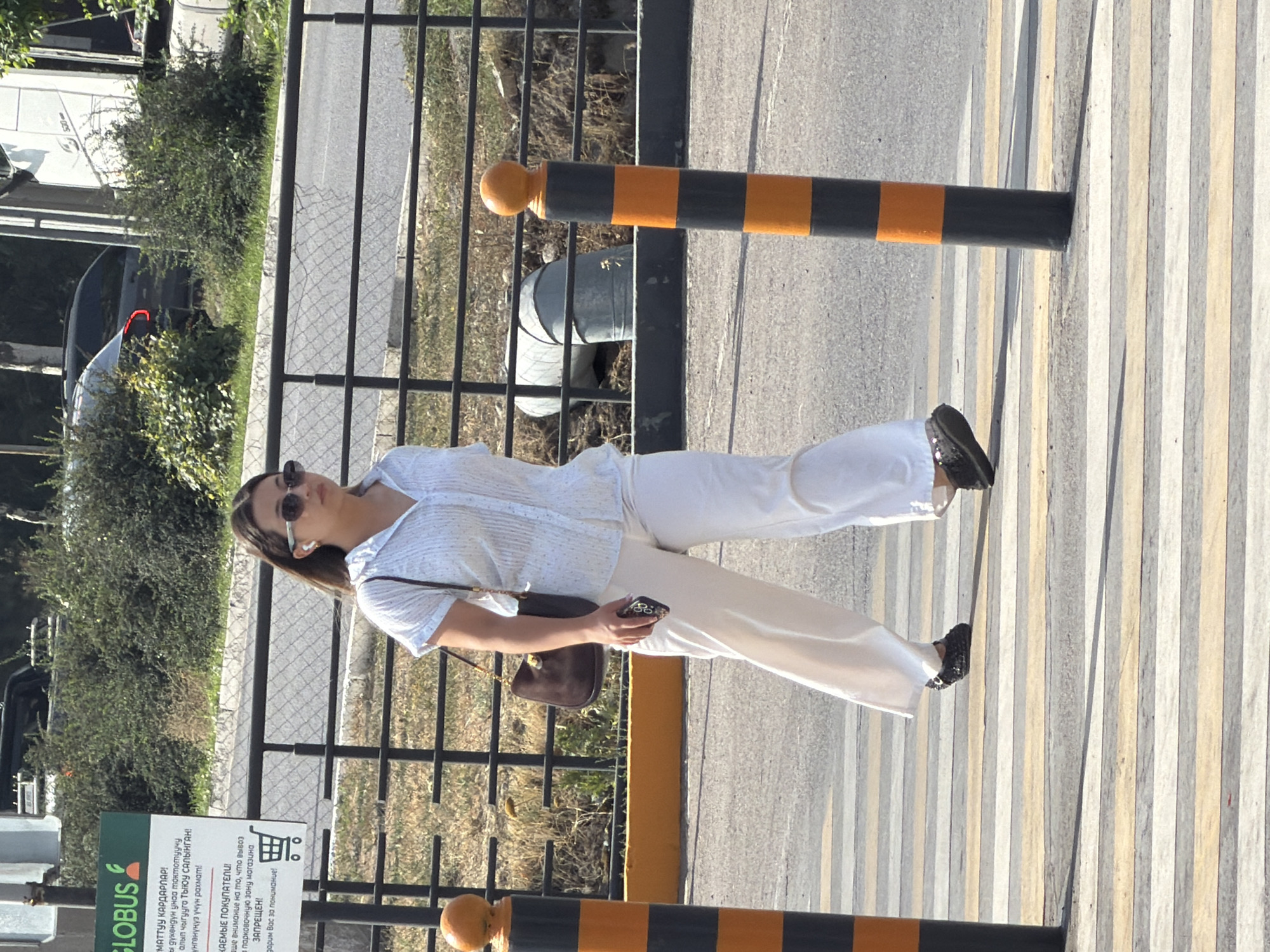 A young woman walks across a crosswalk in a city. She is wearing a light blue patterned short-sleeved shirt, white wide-leg pants, black slip-on shoes, and sunglasses. She has a dark brown shoulder bag and is holding a smartphone in her right hand. In the background, a black metal fence separates the street from a grassy area with bushes. A sign with "GLOBUS" and text in a foreign language is visible on the left. Two orange and black striped bollards are positioned on either side of the crosswalk. A large metal pipe is partially visible in the background behind the fence. The scene is lit by sunlight, casting shadows on the ground.