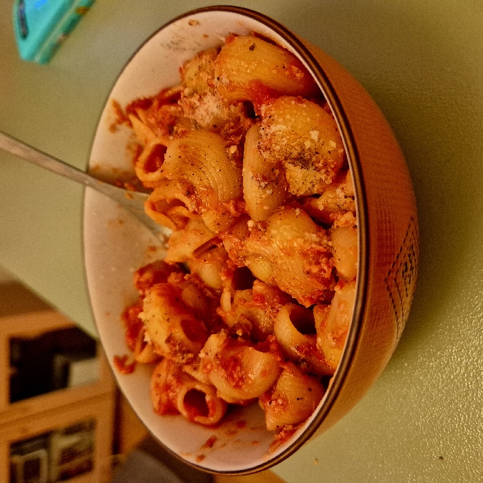 A bowl of pasta, likely lumache or similar shell-shaped pasta, is covered in a red tomato-based sauce and sprinkled with what appears to be Parmesan cheese and black pepper. The pasta is in a white bowl with a brown outer design and rim. A silver fork is visible in the bowl. The bowl sits on a light green surface. A portion of a blue object is present in the upper left corner, and a shelf with a dark object sits in the lower left corner.