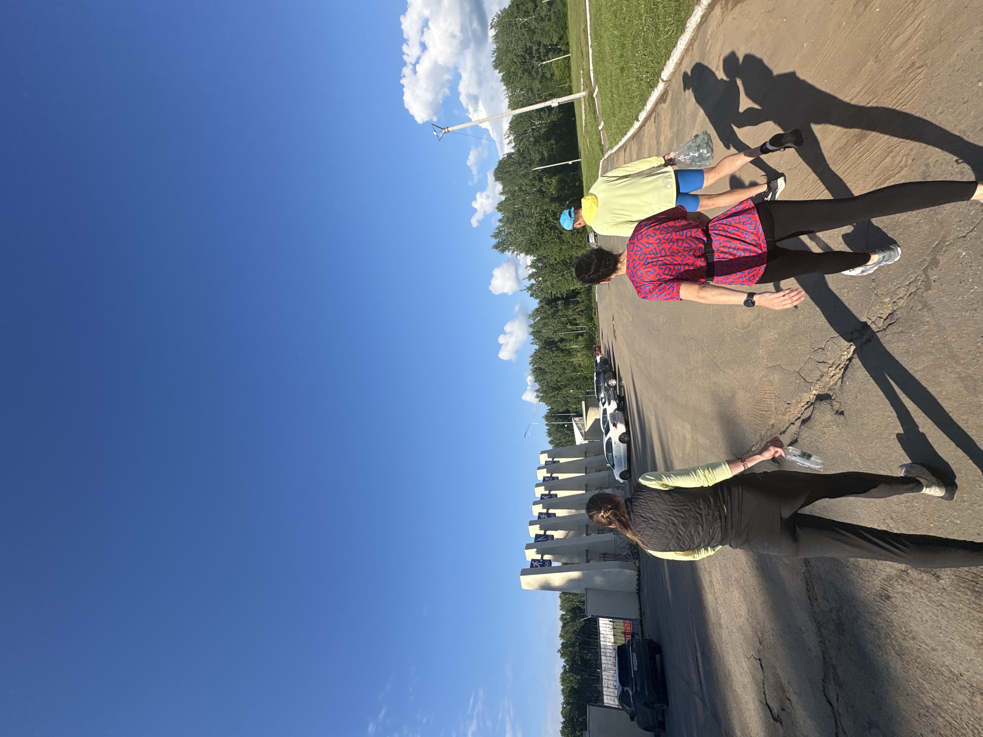 This image captures an outdoor scene in Chaikovsky, Russia, featuring three individuals walking on a paved path during what appears to be late afternoon or early morning due to the long shadows cast by the low sun angle. The weather is clear and sunny, with a bright blue sky dotted with scattered white clouds.
From foreground to background, the first person is seen from behind, wearing a dark vest over a light green long-sleeved shirt, dark pants, and light-colored sneakers, holding a clear water bottle. Ahead of them and slightly to the left, another individual walks in a red and blue patterned short-sleeved shirt, black pants, and grey sneakers, with a watch on their left wrist. The third person, further in the distance, wears a light yellow long-sleeved shirt, blue shorts, and a blue cap, also carrying a water bottle.
The paved path features some cracks and runs alongside a grassy area on the right. A line of lush green trees forms the backdrop along the path. To the left, a series of large, light-colored concrete structures are visible, resembling stadium seating or architectural elements of a sports facility. Several cars, including white and dark models, are parked behind these structures. The scene depicts a casual walk or exercise activity in a recreational setting. No discernible text is visible.
