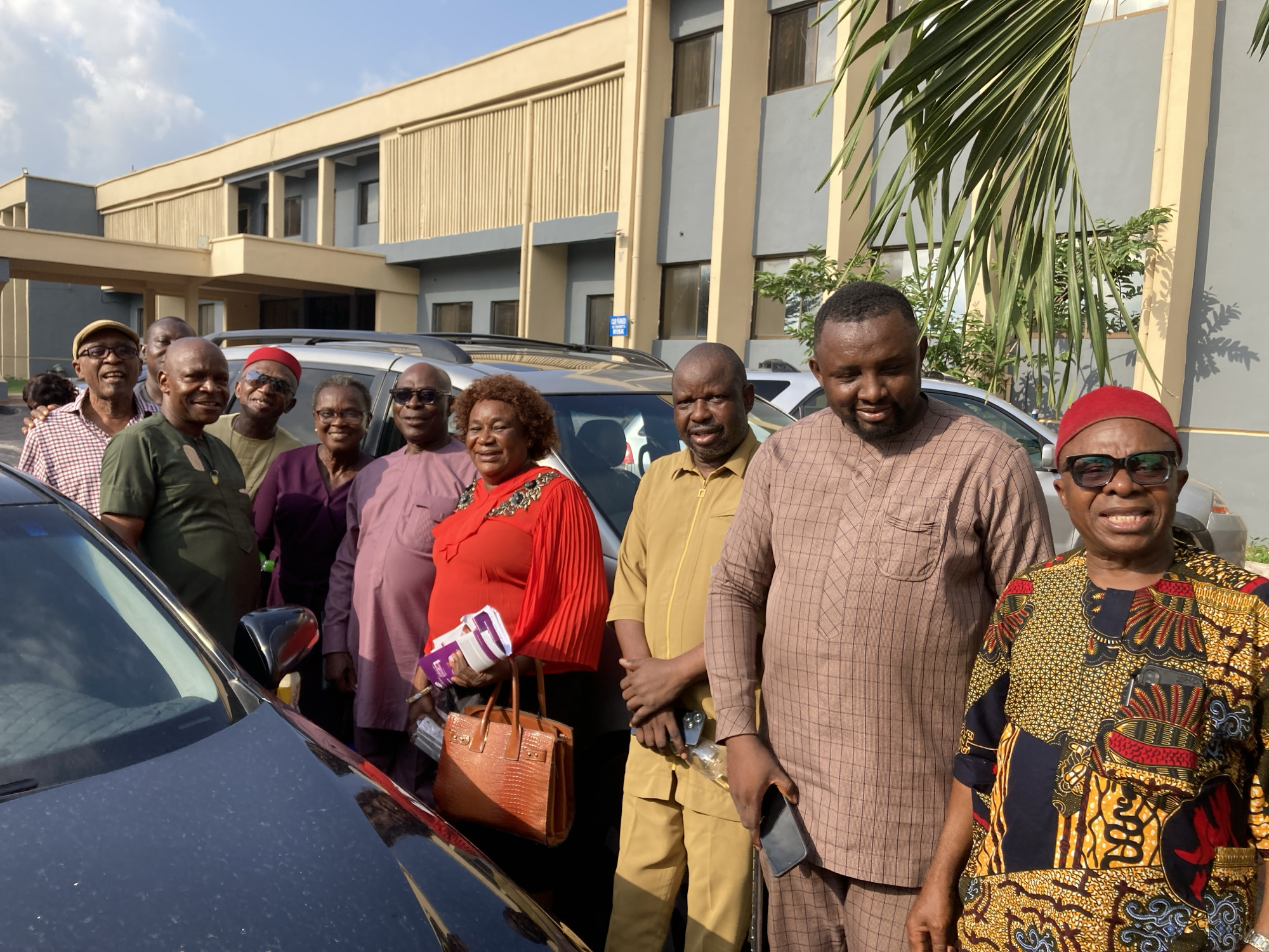 The image features a group of at least ten adults gathered outdoors in what appears to be Umuahia, Nigeria, posing for a photograph. The individuals, a mix of older and younger men and women, are mostly smiling, suggesting a friendly and positive atmosphere.
From left to right, visible subjects include a man in a checkered shirt and cap, a man in a dark green top, a woman in a purple dress and glasses, and a man in a light mauve traditional outfit and glasses. A woman in a bright red top with pleated sleeves holds a brown handbag and a purple item. Next to her is a man in a mustard yellow matching shirt and trousers, holding a phone and a small bottle. He stands beside a man in a brown checkered traditional kaftan, also holding a phone. On the far right, a man in a colorful patterned traditional shirt with a red cap and sunglasses smiles at the camera.
In the background, a modern-looking building with beige and grey facades, multiple windows, and a covered entrance structure is visible. Several vehicles are present; a dark-colored sedan is partially in the foreground on the left, and a silver SUV is parked behind the group. Green palm leaves and other foliage are seen on the right, providing natural elements to the scene. A small blue sign on the building contains the words "CAR PARKED" and "RISK". The bright, natural daylight and clear sky indicate the photo was taken during the day, likely under sunny weather conditions.