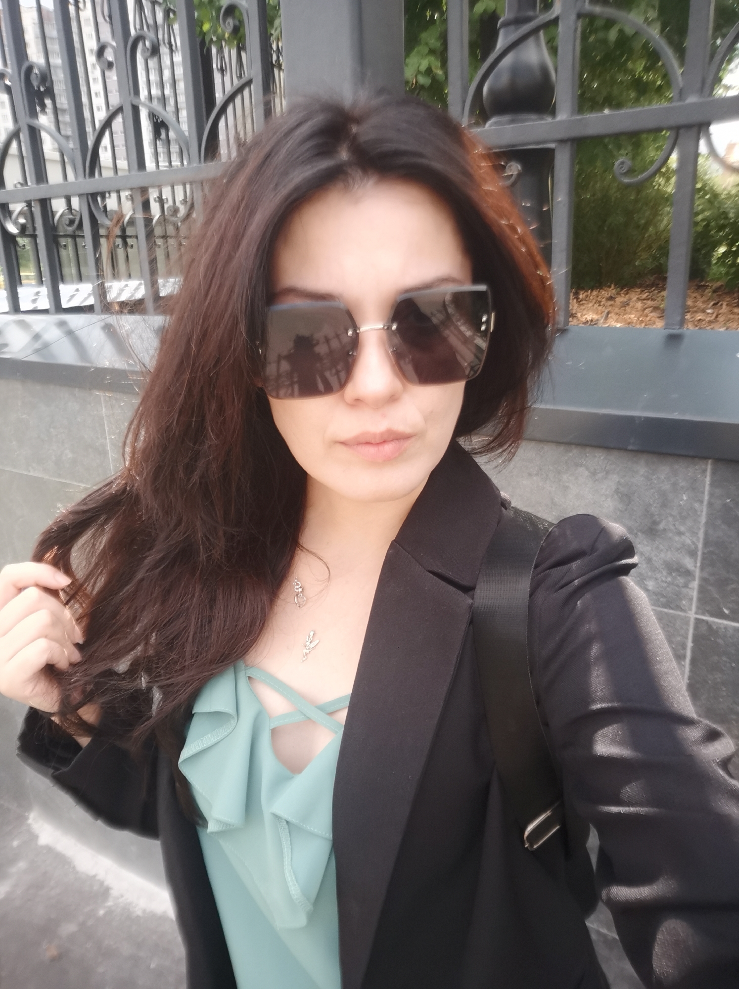 A medium shot, selfie-style photograph shows a woman with long, dark hair and sunglasses posing outdoors. She is wearing a black blazer over a light blue, ruffled top with a criss-cross neckline and a delicate silver necklace. Her left hand is gently holding a strand of her hair. The background features a dark, ornate metal fence, a black lamppost, and a textured wall with stone tiles. Some greenery and buildings are visible behind the fence. The lighting suggests it is daytime, with shadows casting across the scene. The specific location is not discernible from the image itself, making it impossible to confirm if it is in Moscow, Russia.