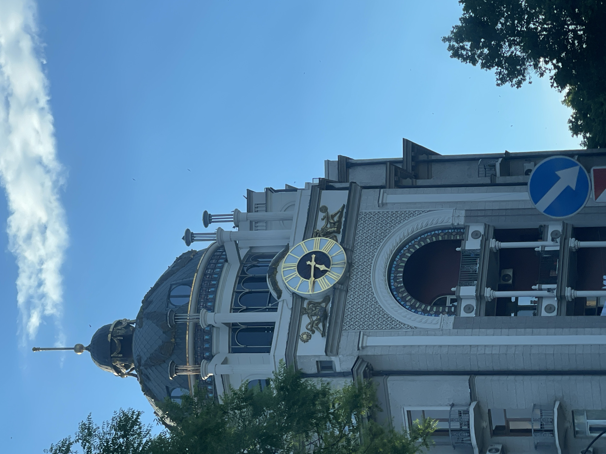 The image depicts the Rezo Gabriadze Theater in Tbilisi, Georgia, from a low angle. The building features a clock tower with a blue-and-gold clock face, an ornate dome with a spire, and a façade with arched windows and decorative elements. A blue directional traffic sign is visible on the right. Trees partially obscure the base of the building and the right side. The sky is clear blue with some wispy clouds.