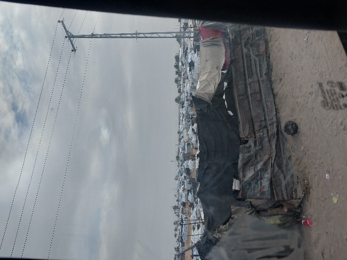 A sprawling settlement of makeshift homes, covered in tarps and corrugated metal, stretches across a flat, dusty landscape. Overhead, power lines run on tall metal towers. The sky is overcast, suggesting a bleak environment. The photograph seems to have been taken from a vehicle. A black object is placed in the foreground which could be part of the vehicle itself.