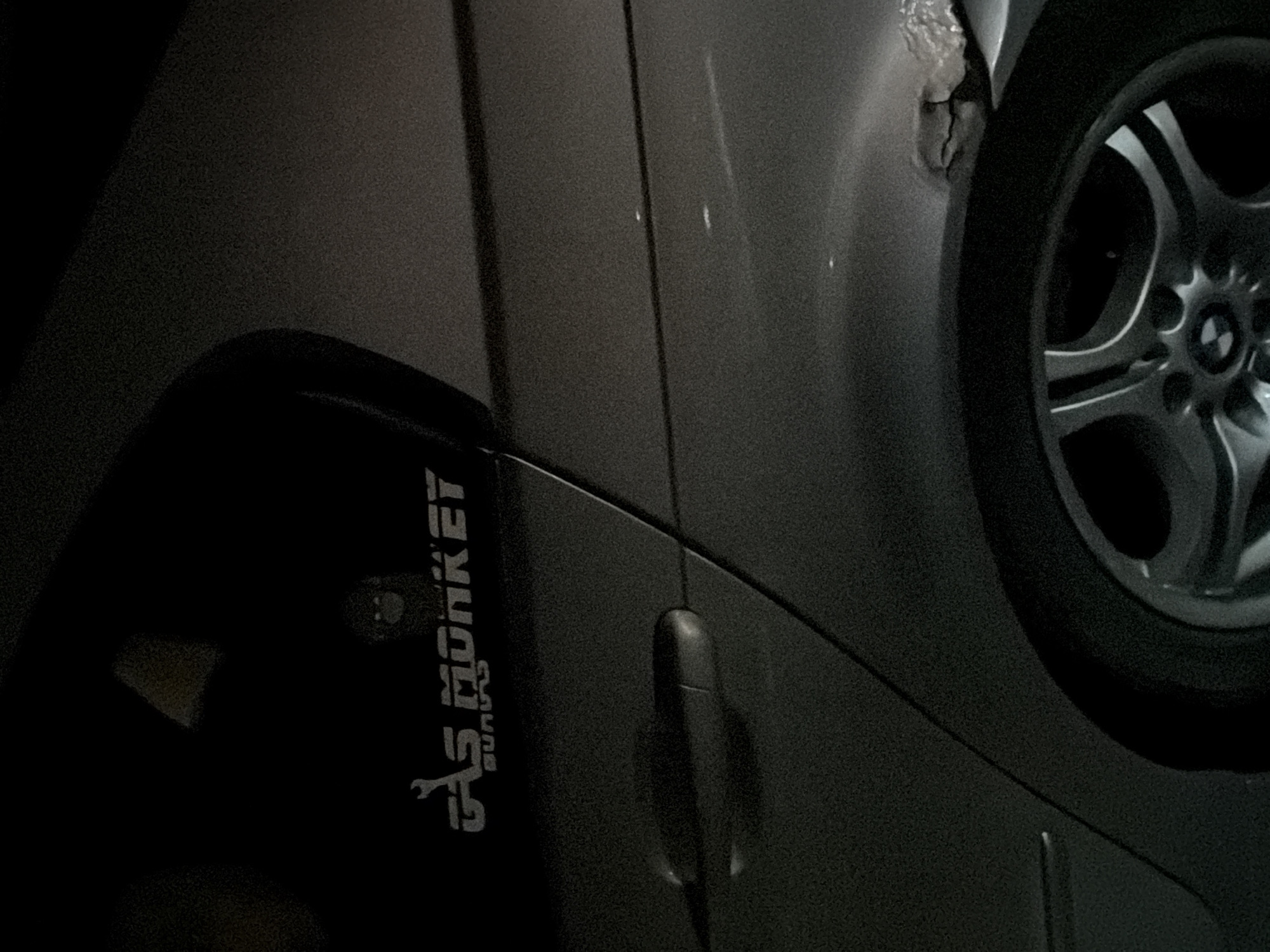 The image shows a portion of a grey car, likely parked on a street in Nesebar, Bulgaria. A "Gas Monkey Garage" sticker is visible on the rear door. The car has a door handle and a section of the rear wheel. There is visible rust and damage to the car's bodywork near the wheel arch. The lighting suggests it is either nighttime or the area is poorly lit. No people are present in the frame.