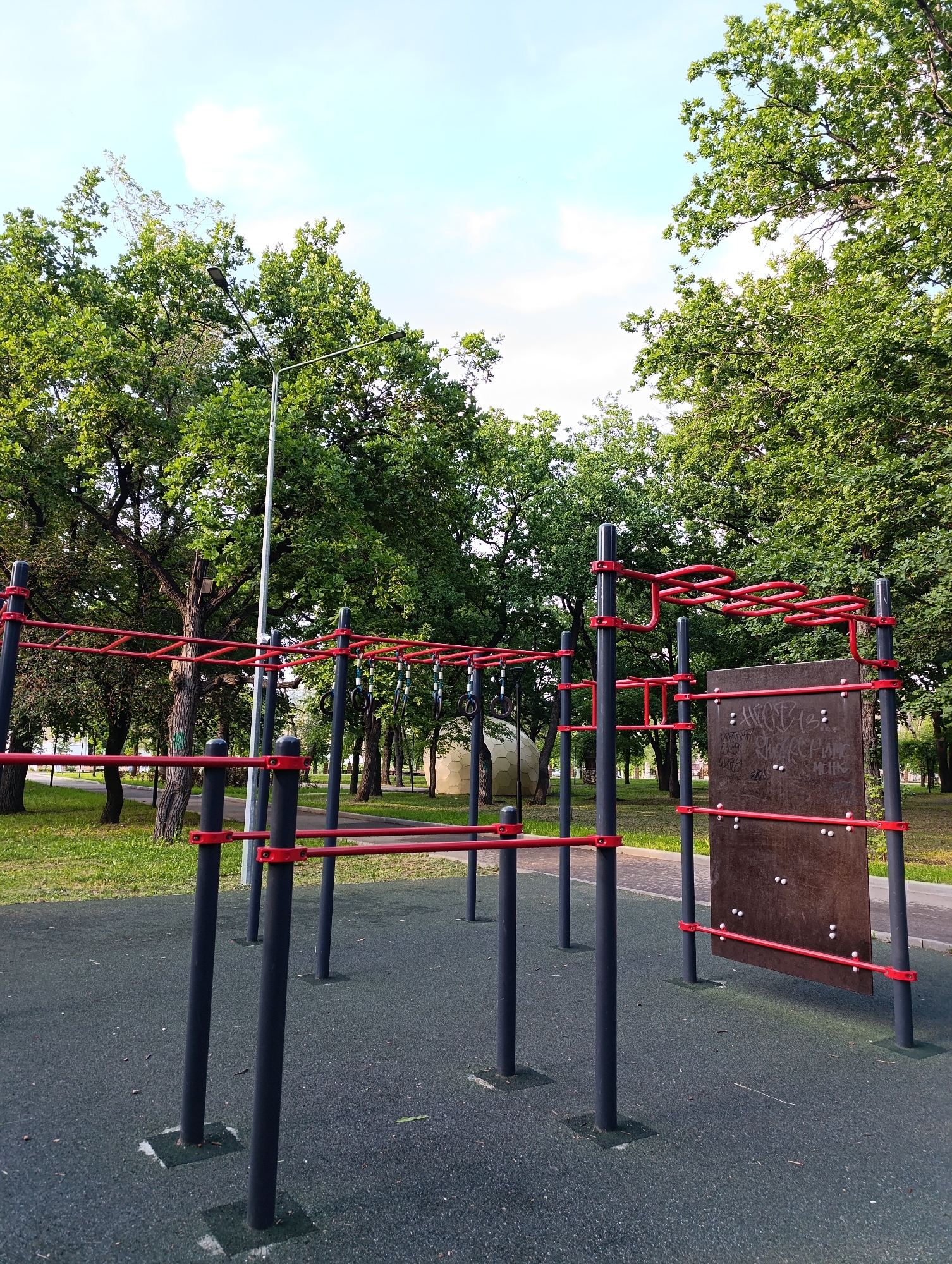 A low-angle shot shows an outdoor calisthenics gym in Novokuybyshevsk, Russia. The gym features several metal bars and posts, painted black with red accents. A climbing structure with a zig-zagging red bar is prominent in the background. To the right, a large wooden board is attached to the structure, covered in graffiti. The ground is a dark, textured rubber surface. Lush green trees with full canopies surround the gym, suggesting a park or recreational area. A lamppost stands in the middle ground. The sky is visible above, with scattered clouds against a pale blue backdrop, indicating daytime. There are no people present in the image. The scene appears peaceful and ready for use, with the time of day likely late afternoon or early evening based on the light. The graffiti on the wooden board includes Cyrillic lettering, but the text is not clearly legible.