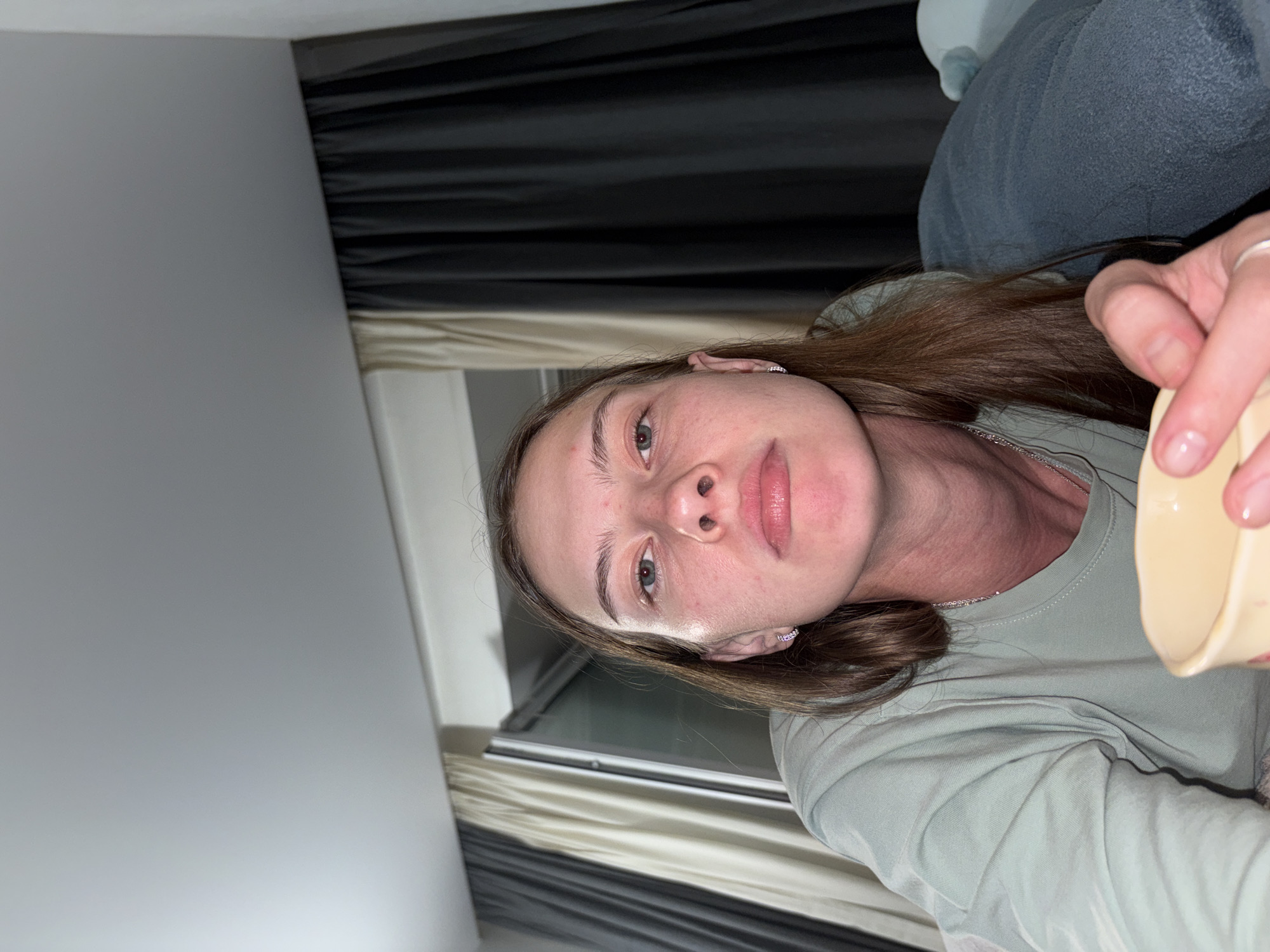 The image shows a medium shot of a woman lying down, possibly in a bedroom. She is looking directly at the camera with a neutral expression. She has long brown hair, light skin, and appears to be wearing a light green long-sleeved shirt. She is holding a light yellow mug in her right hand. In the background, there are curtains, with one set appearing to be a light beige and the other a dark grey. There is also a window visible behind the curtains. The lighting suggests it is indoors and possibly evening or a dimly lit room. No specific location in the city of Perm, Russia can be identified from the image.
