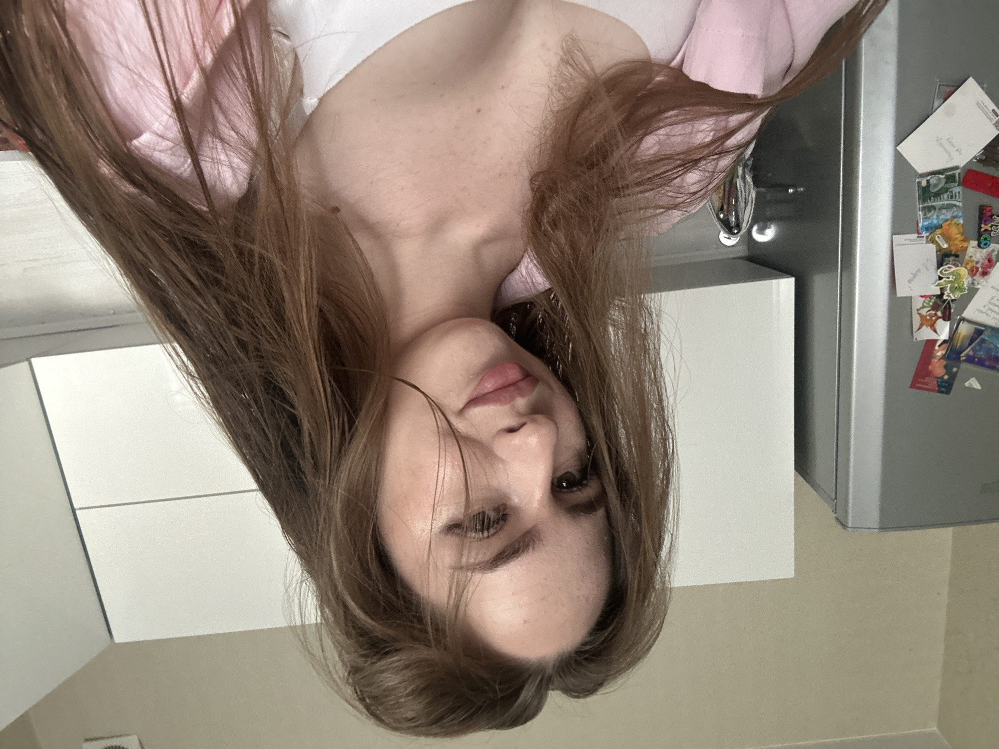 A woman with long brown hair is pictured from the chest up, facing the camera at a slight angle. She is wearing a pink collared shirt open over a white top. Her hair is loose and falls around her shoulders and chest. The background shows a light-colored wall and cabinetry, with the side of a silver refrigerator visible on the left. The refrigerator has several colorful stickers and papers attached to it. The lighting appears to be indoor, with a soft light source illuminating the scene.