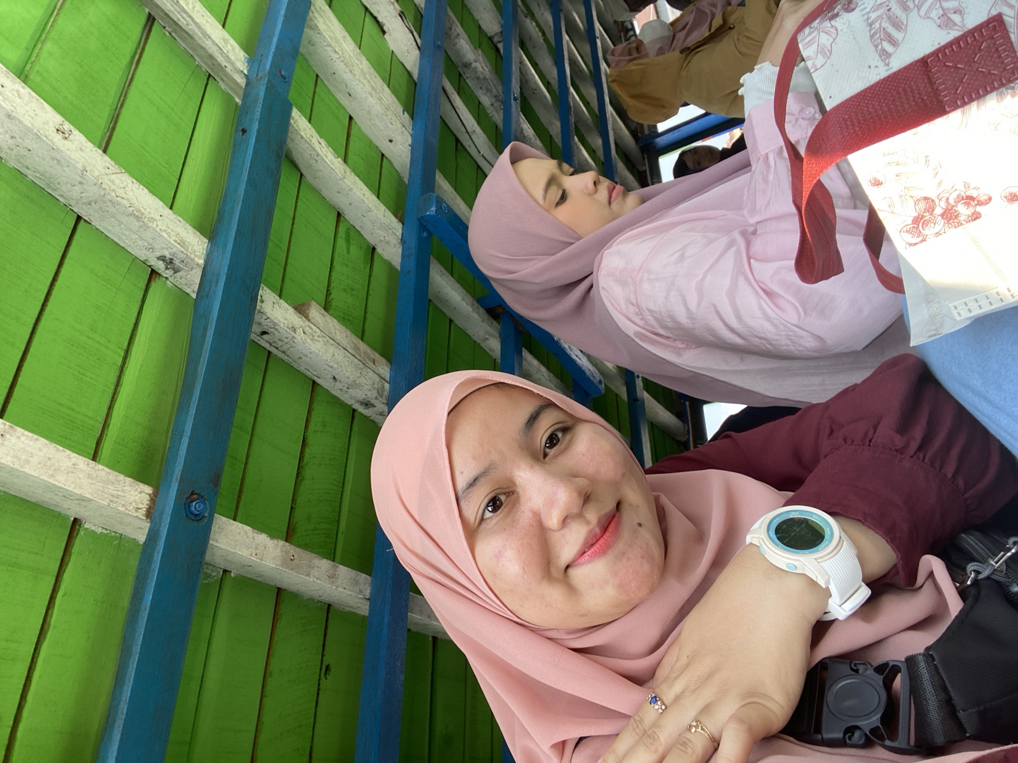The image shows three women wearing hijabs inside what appears to be a covered boat. The woman in the foreground is looking at the camera and smiling. She is wearing a pink hijab and a maroon top, and a white digital watch. She has jewelry on her finger. Another woman in a light pink hijab and shirt is to her right, looking down. Further back, a woman in a yellow hooded jacket and a pink hijab is visible, as well as another person partially obscured. The ceiling of the boat is made of green wooden planks with blue structural beams and white supports. The environment suggests an outdoor setting, possibly on a waterway, characteristic of Banjarmasin, Indonesia, known for its riverine landscape and floating markets.
