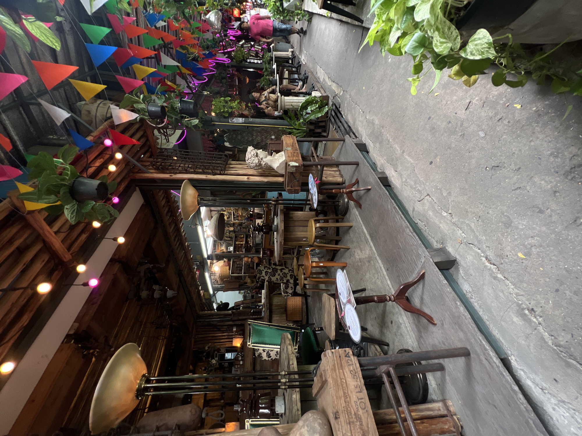 The image depicts a narrow, cluttered alleyway in Bangkok, Thailand, possibly a market or retail area. Colorful triangular flags and hanging plants decorate the overhead space, casting shadows and adding a festive atmosphere. Strings of lights, some with colored bulbs, are strung along the wooden roof structure on the left.
The alley floor is concrete, with a visible seam or channel running along its length. Various objects, including wooden tables, chairs, stools, and vintage items, are placed on the concrete surface. These items appear to be part of a shop or display, filling the narrow space.
A couple of people are visible in the distance, walking through the alleyway. In the foreground, a large wooden box with printed text and numbers is prominent. The overall scene is busy, eclectic, and characteristic of some market areas in Bangkok.