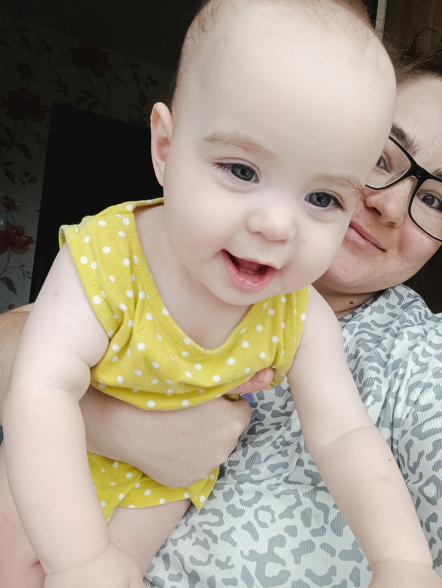 A close-up, medium shot captures a baby being held by an adult. The baby, in the foreground, is wearing a yellow sleeveless top with white polka dots. The baby's face is the primary focus, with a wide, happy smile and blue eyes looking towards the viewer. The adult, partially visible behind the baby, is wearing glasses and a patterned shirt. They are looking at the camera. The background shows a dark wall with floral wallpaper and a dark rectangular object, possibly a door or a piece of furniture. There is no identifiable information about the location in Noviy, Russia.