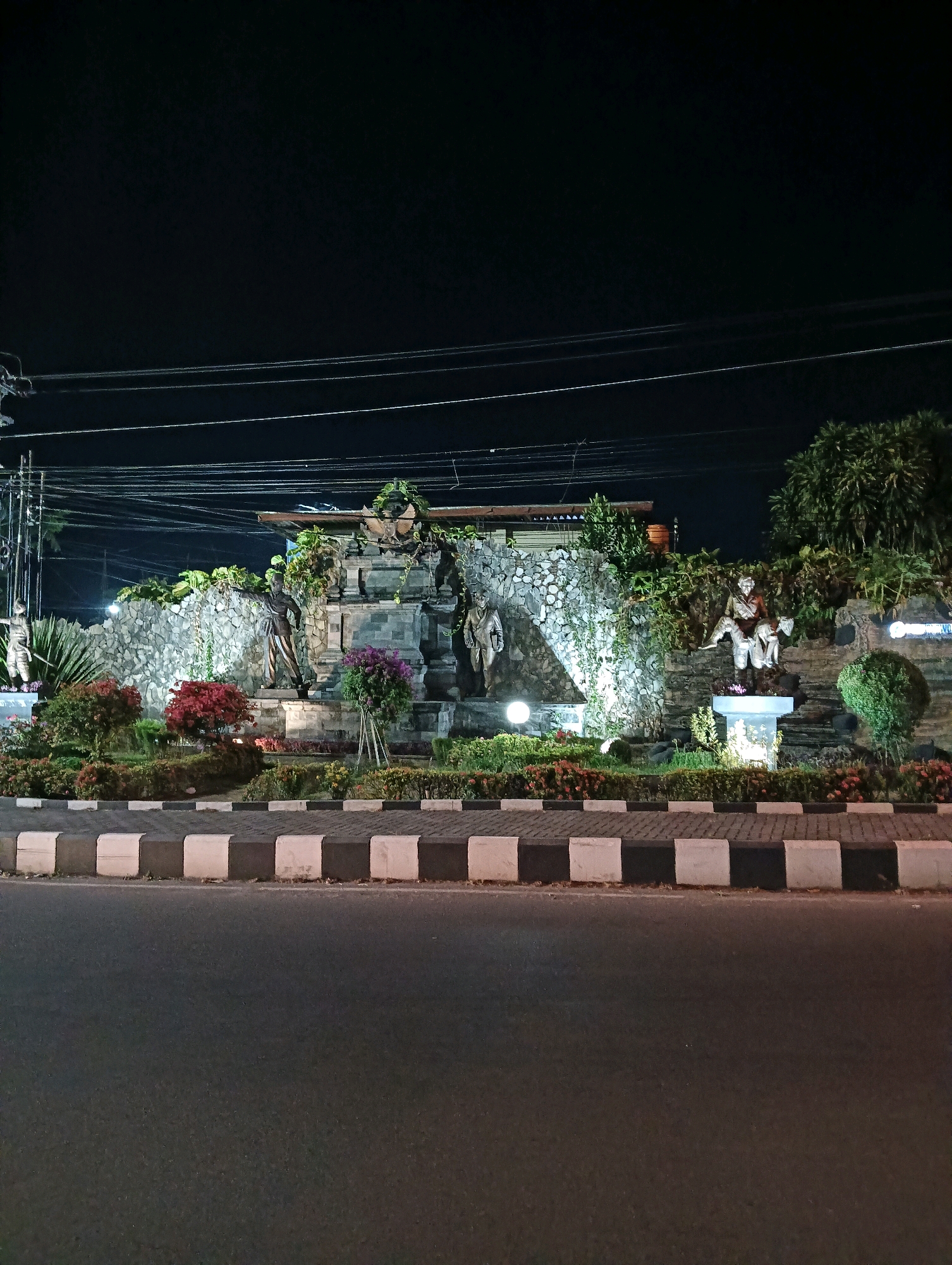 The image shows a roundabout in the city of Purworejo, Indonesia, at night. A stone monument with sculptures and lush greenery is illuminated by spotlights. Several bronze statues of figures in traditional attire are displayed on and around the monument. The roundabout is bordered by a pedestrian walkway with black and white striped curbing, and a paved road is visible in the foreground. Overhead, power lines stretch across the dark sky.