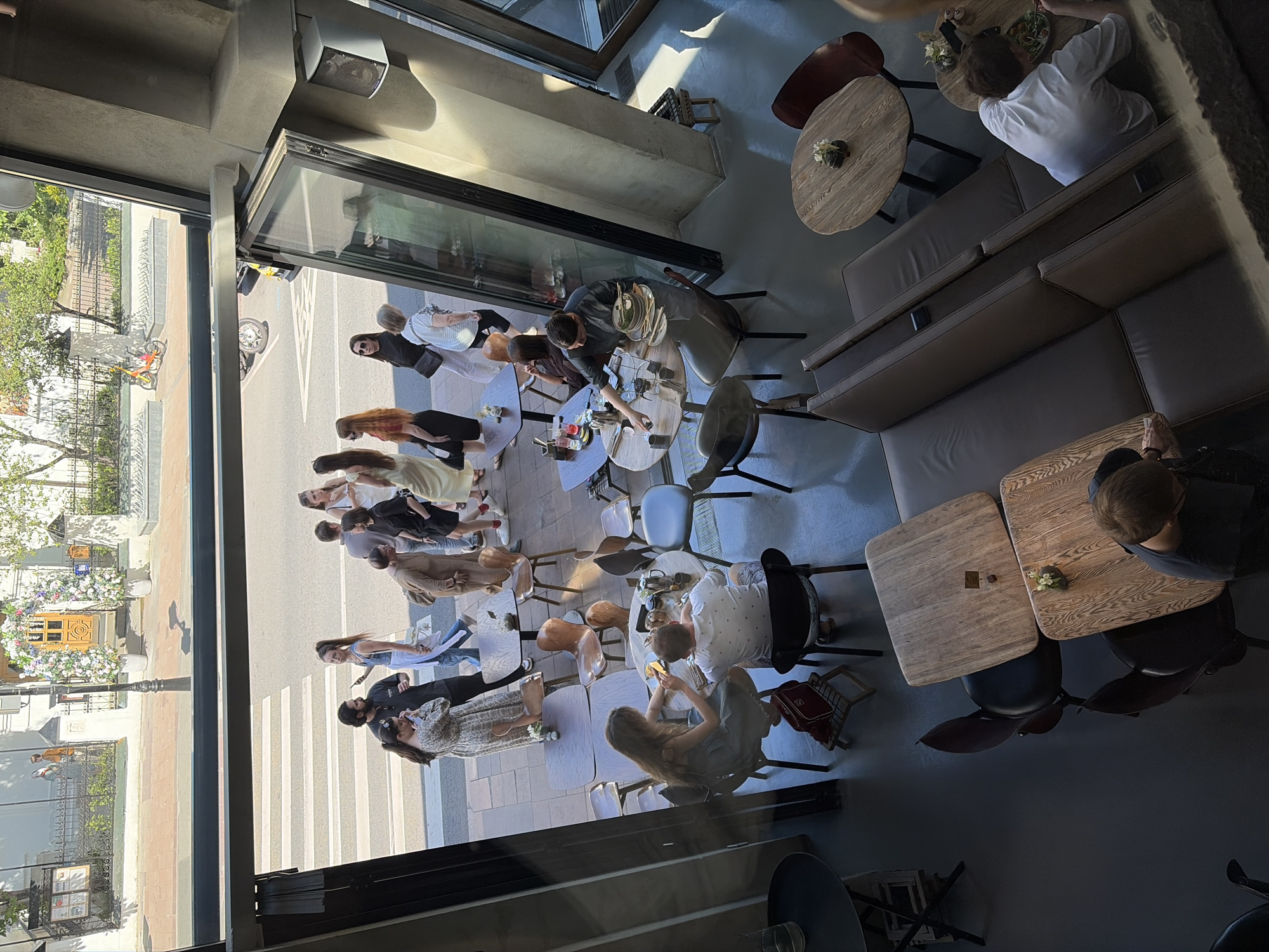 The image captures an interior view from above, looking out onto a street and into a cafe. Outside, people are walking across a crosswalk and standing on the sidewalk. A yellow bicycle is parked near a building with decorative floral arches and a wooden door. Inside the cafe, patrons are seated at various tables, some eating, some talking, and some looking at their phones. The cafe has modern furniture, including wooden tables and chairs, as well as some plush seating. The overall scene depicts a vibrant urban environment on a sunny day.