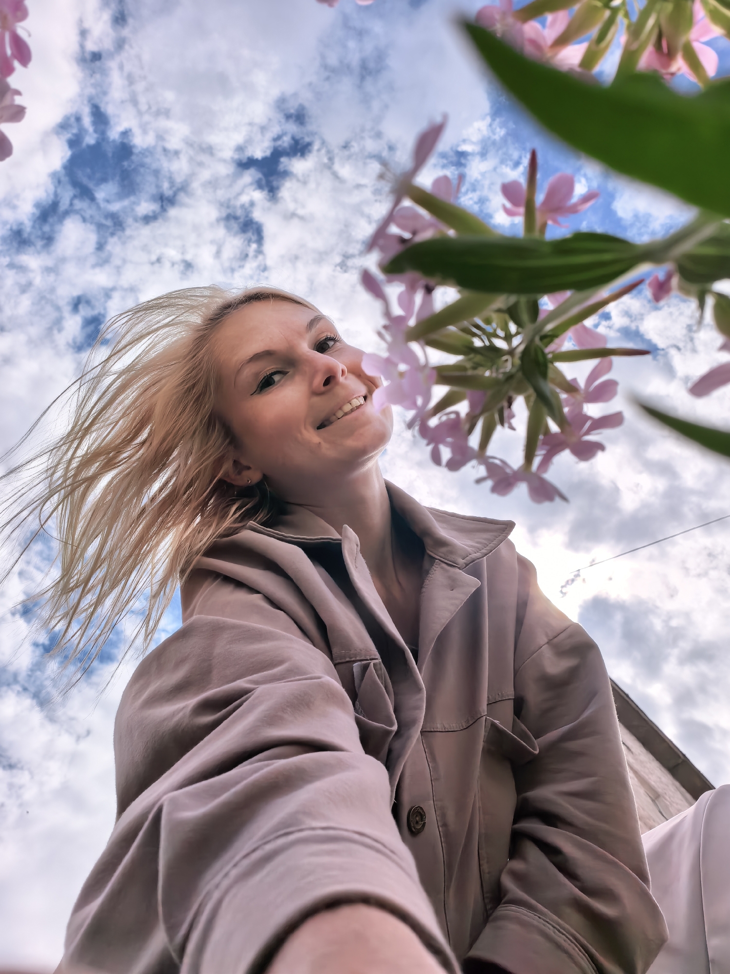A low-angle shot captures a smiling young woman from the chest up, looking upwards towards the camera. Her blonde hair, appearing windblown, frames her face. She is wearing a light brown or tan collared jacket. Behind her, a blue sky with fluffy white clouds is visible, with glimpses of pink flowers and green leaves in the foreground, suggesting an outdoor setting during the daytime. The overall mood is cheerful and bright, possibly indicating a pleasant day. The image was taken in Sterlitamak, Russia. There is no visible text in the image.