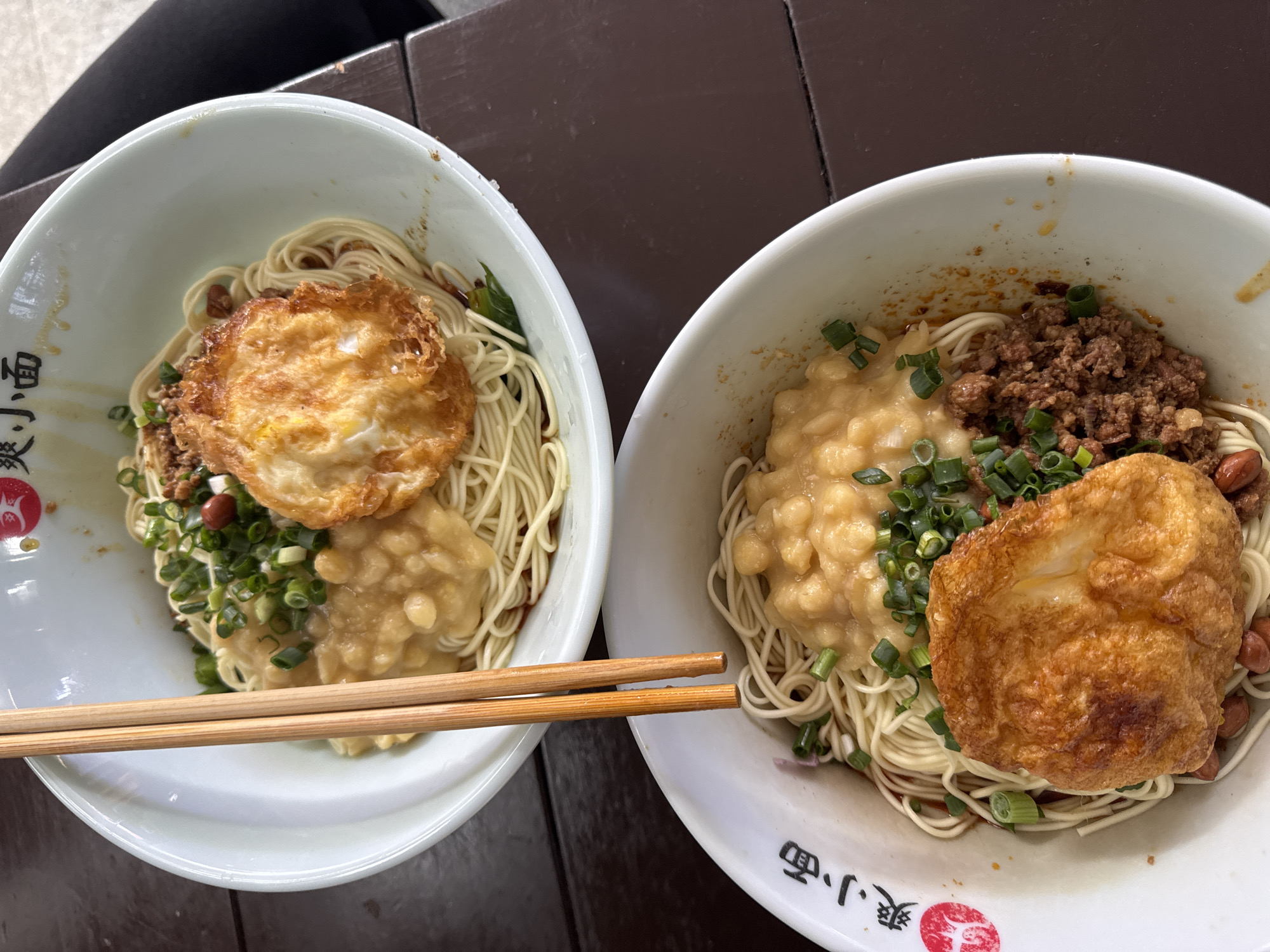 Two bowls of noodles are placed on a dark brown wooden surface. The bowl on the left has noodles topped with a fried egg, a dollop of pale yellow sauce with small white chunks, and sprinkled green onions. Wooden chopsticks rest on the bowl's rim. The bowl on the right contains noodles topped with a fried egg, ground meat, a dollop of pale yellow sauce, sprinkled green onions, and a few peanuts. The bowls are white with Chinese characters printed on the rim.
Based on the cuisine, presence of Chinese characters, and details of the environment, this image likely originates from Yuzhong District, China.