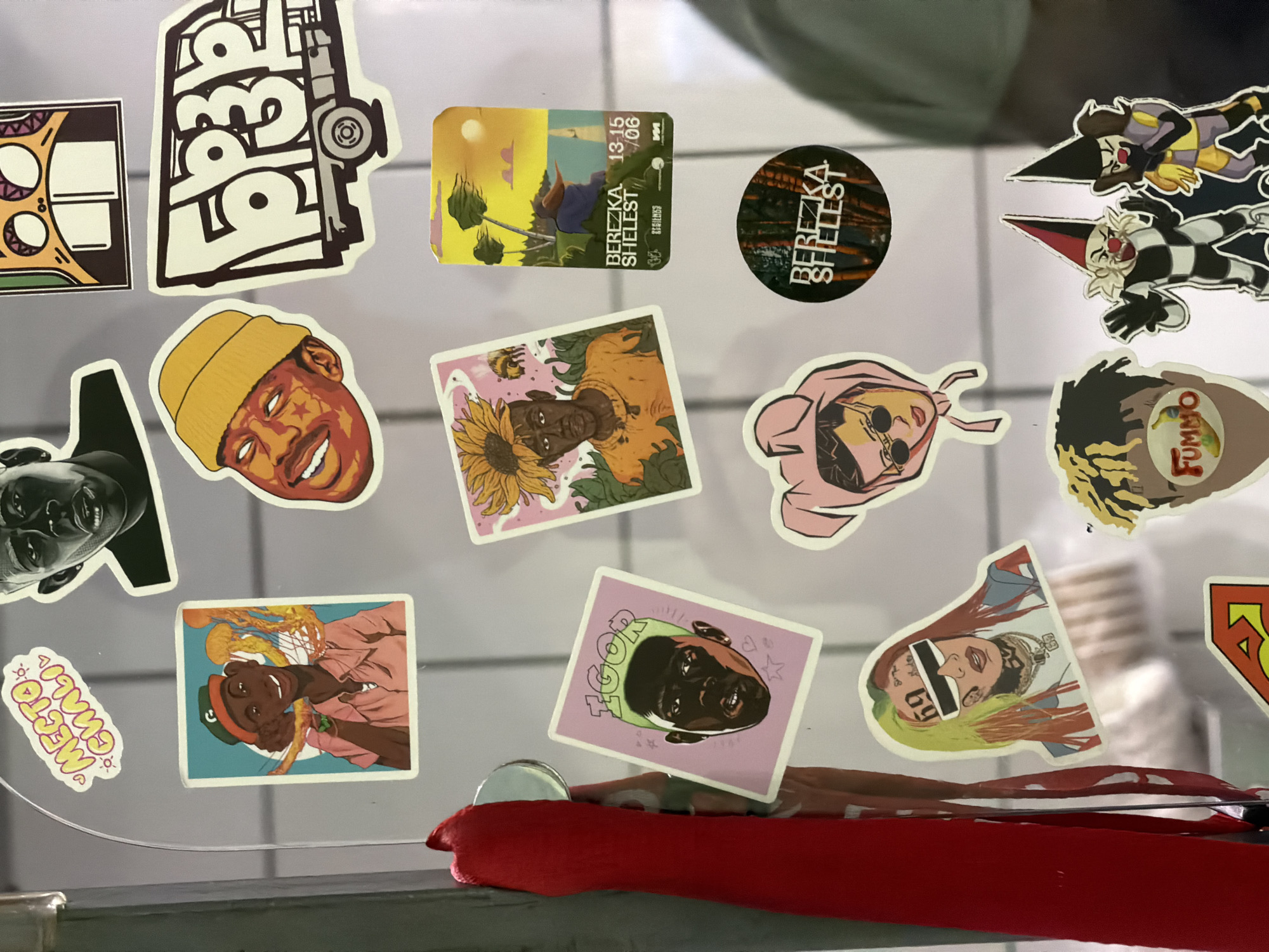 The image shows a collection of stickers on a refrigerator door. The stickers feature various cartoonish illustrations, some of which appear to be inspired by hip-hop culture, including depictions of rappers and album art. One sticker shows a character with the name "IGOR" and another with "69" and sunglasses. There are also stickers with text in Russian, such as "MECTO CUAH" and "БЕРЕЗКА SHELEST". The refrigerator door is white and a red lanyard with a metallic object is attached to the side. The background is blurred but appears to be an indoor environment. There is no discernible information that places the scene specifically in Moscow, Russia.