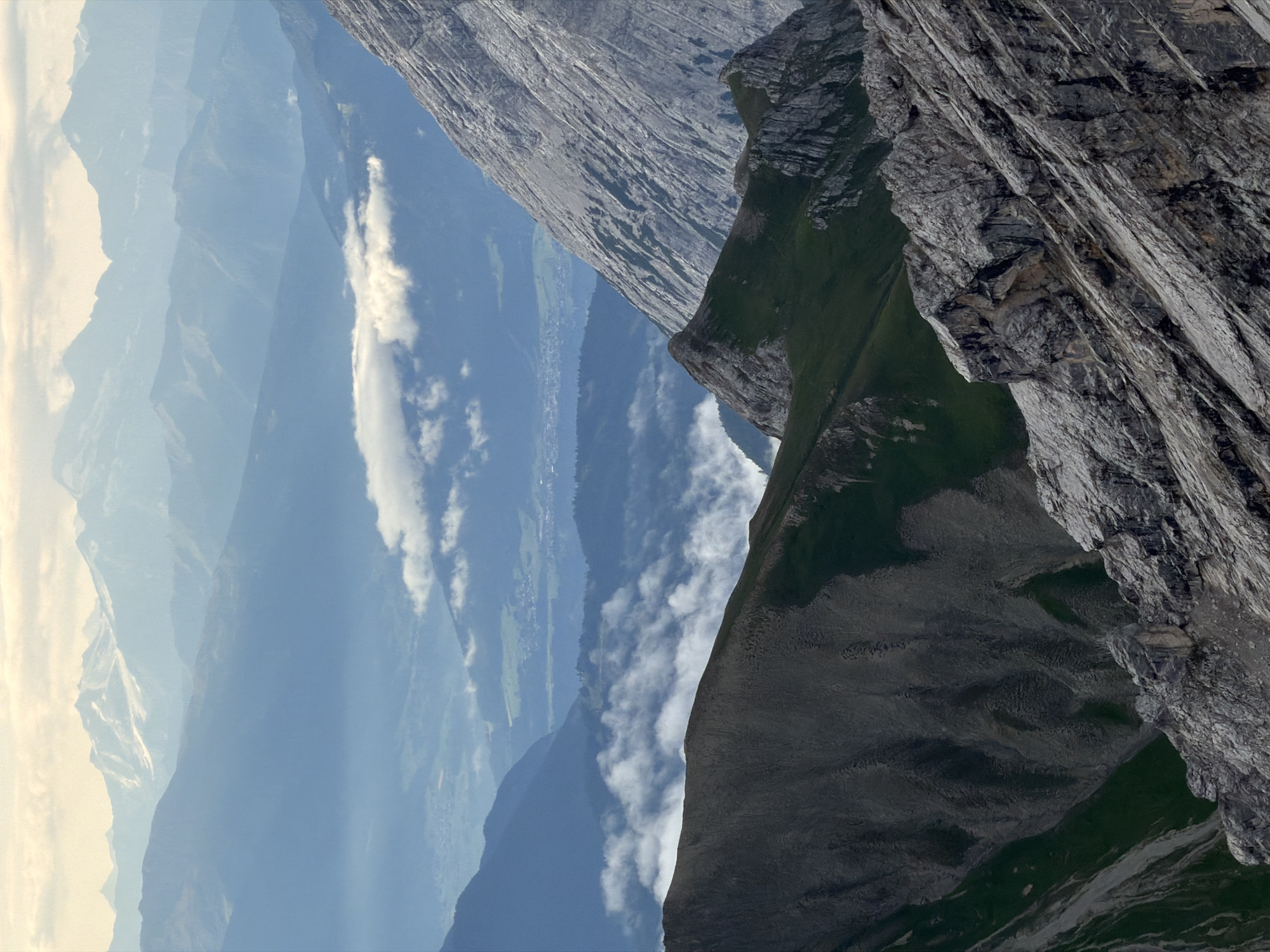 The image depicts a majestic mountain landscape with multiple layers of peaks receding into the distance. The foreground is dominated by rugged, rocky slopes, with one particularly prominent, steep, gray mountain face displaying distinct striations and textured rock formations. To the left and in the mid-ground, there are dark, shadowy mountain slopes with patches of green vegetation. Wisps of white clouds are scattered throughout the valleys and between the mountain ranges, adding depth and atmosphere. In the far distance, hazy, blue-hued mountains are visible, some with snow-capped peaks, under a pale, overcast sky. A faint, distant valley with some signs of human habitation, likely a town or village, can be discerned amidst the haze. The overall impression is one of a vast, natural, and somewhat dramatic alpine environment. The location is likely in the vicinity of Garmisch-Partenkirchen, Germany, a region known for its stunning mountain scenery. There are no people or discernible human activity in the image.