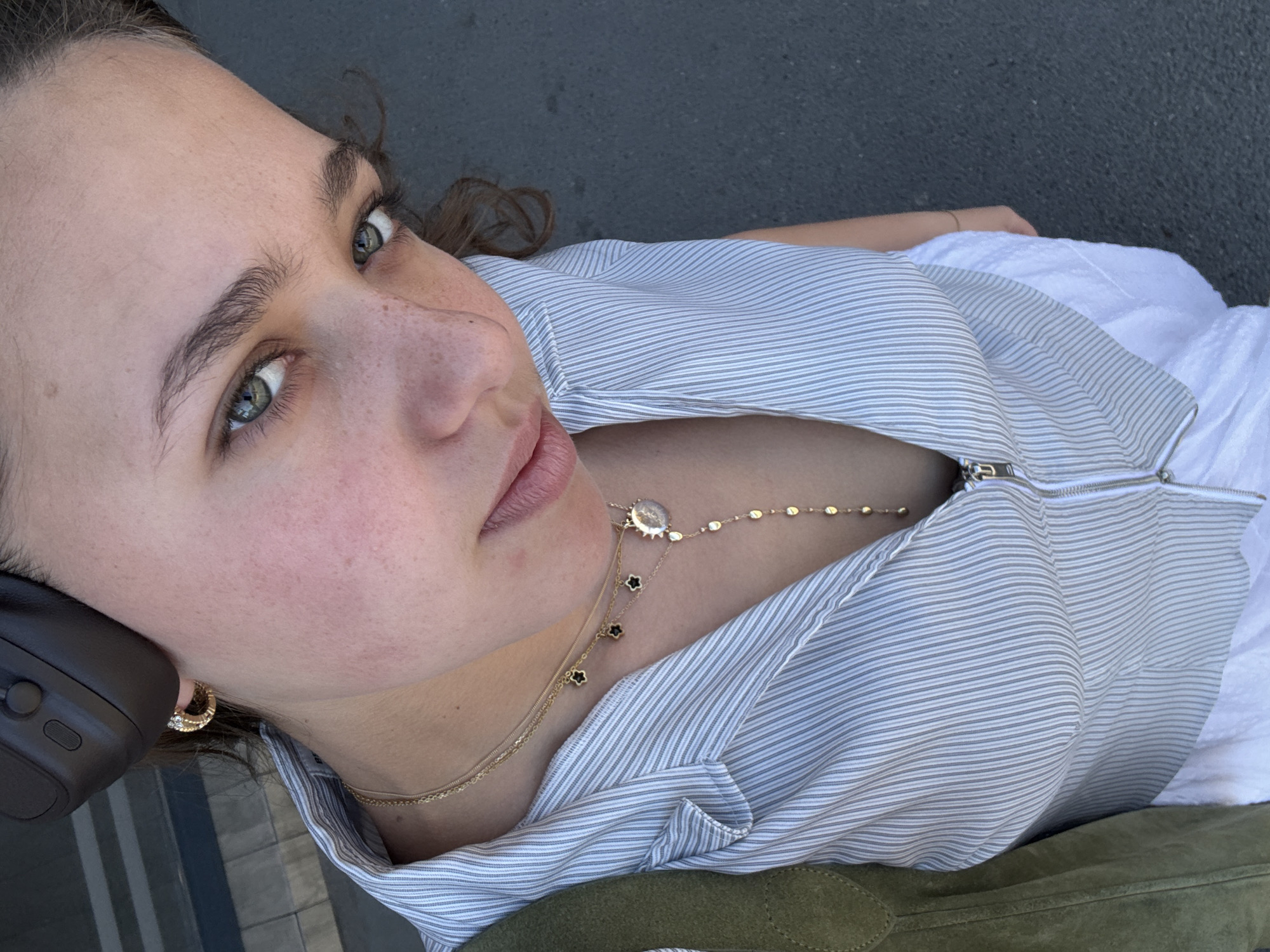 A close-up, angled shot captures a woman with fair skin and green eyes. She wears a horizontally striped gray and white top, unzipped at the collar, revealing layered gold necklaces. Her attire is complemented by a white garment beneath the top and an olive green jacket or accessory visible in the lower portion of the frame. Gold hoop earrings adorn her ears. The scene suggests an outdoor urban setting. The location of the photo is unknown.