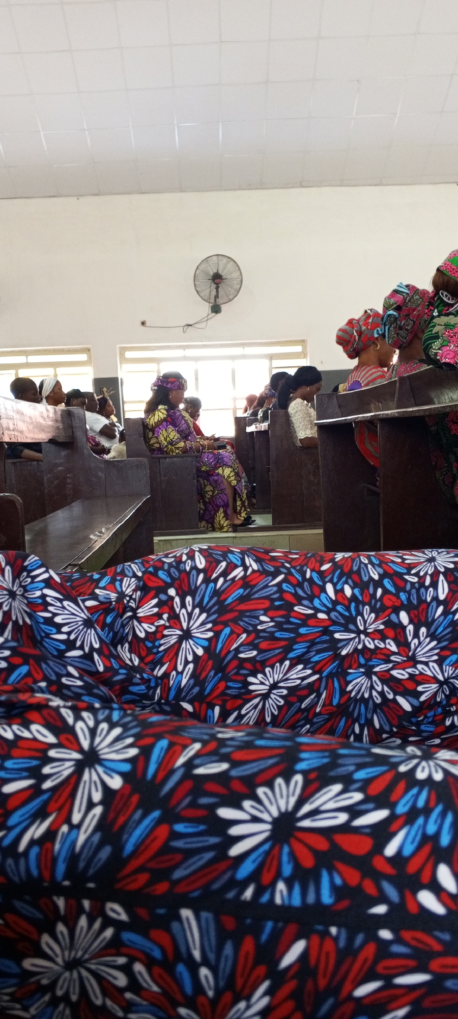 In an indoor setting that appears to be a church hall in Jalingo, Nigeria, several individuals are seated on wooden pews. The room has a white tiled ceiling and cream-colored walls, with windows visible in the background. A black oscillating fan is mounted high on the wall. The foreground is dominated by a folded fabric with a vibrant, abstract floral pattern in red, white, and blue.
In the mid-ground, various people are seated, mostly women, facing towards the front of the room. One woman in the foreground, seated to the left of center, is wearing a colorful dress with a yellow and purple floral print and a matching headscarf. She is looking down, possibly at a book or her phone. To her right, other individuals are visible, some with headscarves in various colors and patterns, suggesting a gathering or service. The wooden pews are worn, indicating their age and use. The overall atmosphere appears to be that of a community gathering.