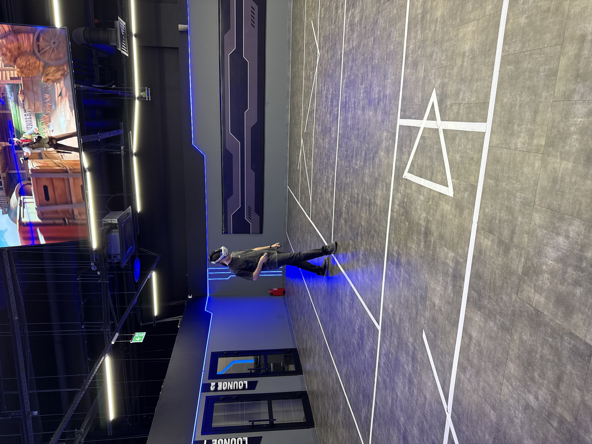 A medium indoor shot reveals a man in a virtual reality setup, standing in a room that appears to be a VR gaming center in the city of Перовское сельское поселение, Ukraine. The room has a dark gray floor with white taped lines that mark out playing boundaries, and a large white triangle shape painted in one of the areas. The walls are a mix of dark gray and black, with blue neon light accents outlining panels and doorways. The man is facing to the right wearing a dark t-shirt, dark pants, and a VR headset. There is a large screen to the left displaying a video game. The lighting in the room is a mixture of fluorescent tube lighting and colored neon accents, contributing to a futuristic ambiance. The words "LOUNGE 1" and "LOUNGE 2" are visible on doors behind the man. A red fire extinguisher is mounted on the wall behind the man.