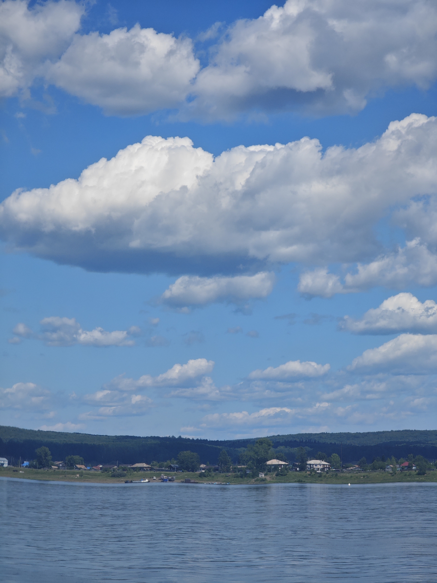 The image depicts a wide, tranquil body of water in the foreground, with gentle ripples on its surface. Across the water, a verdant shoreline is visible, characterized by a dense line of trees forming a natural backdrop. Nestled along the shoreline is a small village or settlement, indicated by the presence of several houses, some with visible roofs and windows. A few boats are also present, moored near the shore, suggesting a connection to the water for transport or fishing. The sky above is a vibrant blue, adorned with numerous fluffy white clouds of varying sizes and densities, creating a picturesque and serene atmosphere. The scene appears to be set in a natural, rural environment, possibly a lakeside or riverbank in Strelka, Russia.