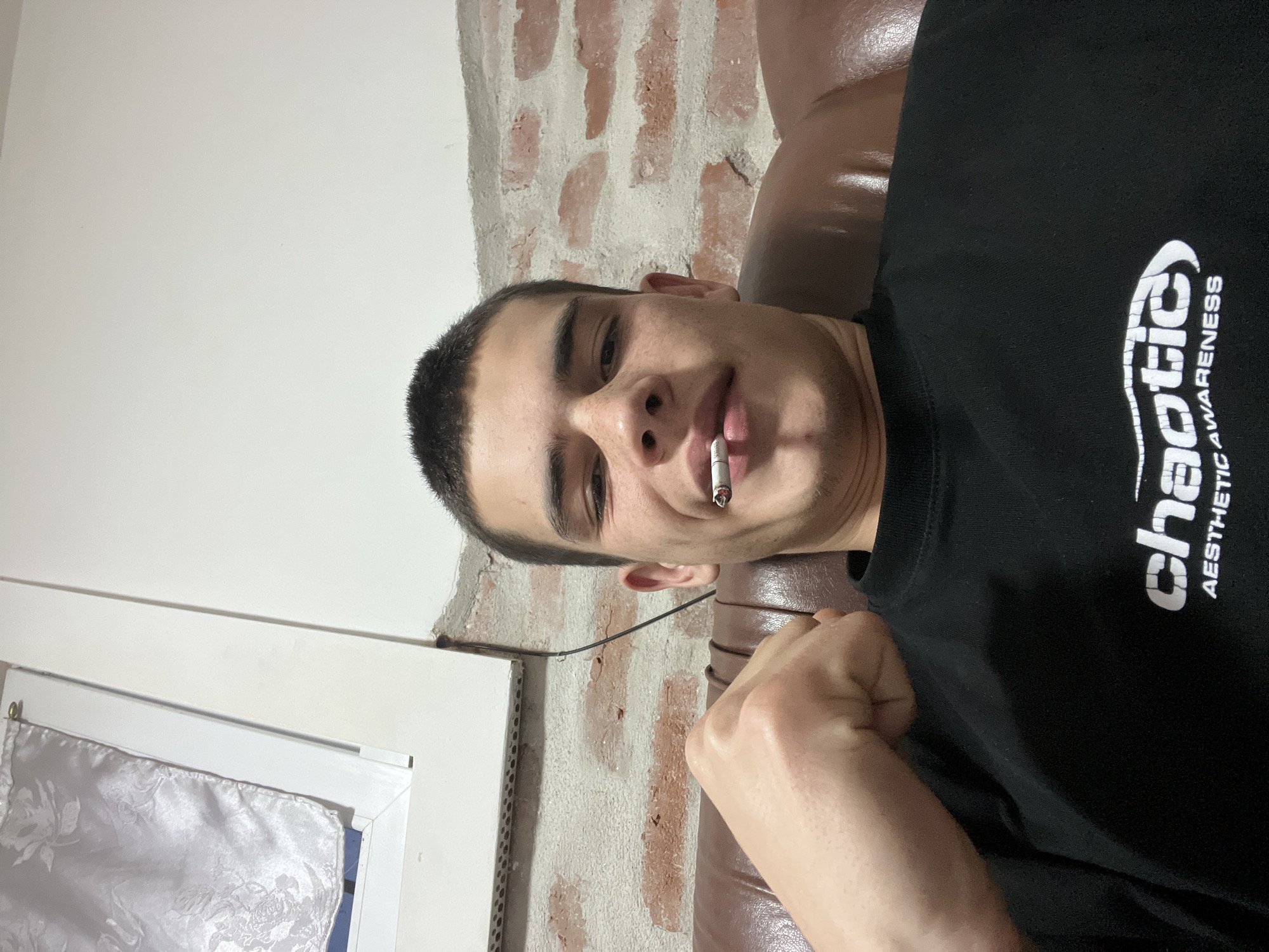 A young man is lying on a brown leather couch, holding a cigarette in his mouth. He is wearing a black t-shirt with the word "chaotic" and "AESTHETIC AWARENESS" printed in white. The background includes a partially brick wall and a white wall with a window frame. The location is not specified.