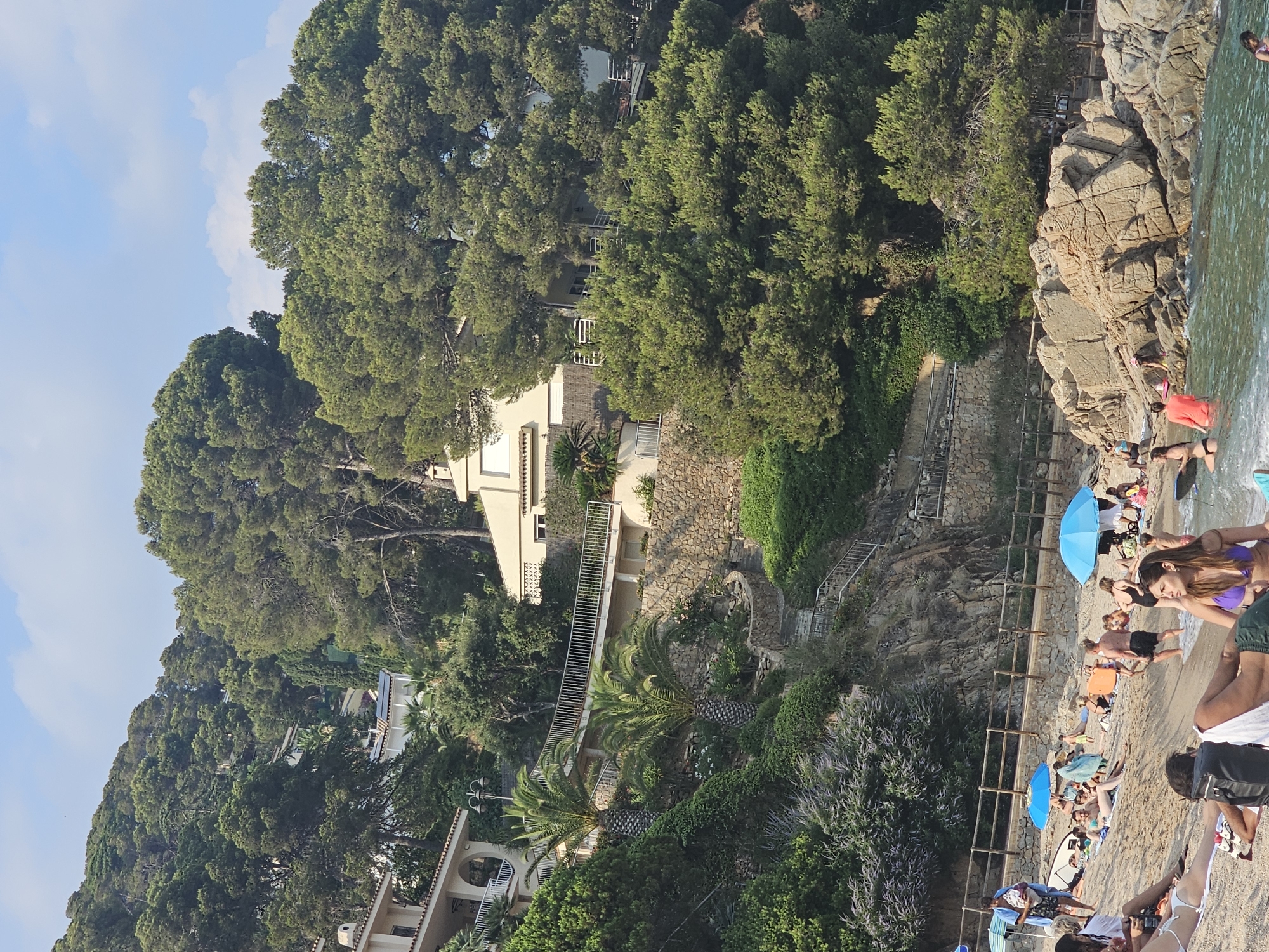 This media file depicts a vibrant beach scene in Blanes, Spain, bathed in the warm light of what appears to be late afternoon or early evening, given the soft shadows and the golden hue on the sand and water. The sky is a clear blue with scattered, wispy clouds, suggesting pleasant weather.
The scene is a rocky coastline bordered by lush green pine trees that cascade down the hillside. Several buildings, typical of Mediterranean architecture with white facades and terracotta roofs, are nestled amongst the trees, suggesting a residential or resort area. Stone pathways and metal railings wind through the landscape, indicating access to the beach.
The beach itself is a sandy strip populated by numerous people enjoying a day by the sea. They are engaged in typical beach activities: sunbathing on towels and loungers, some are in the water, and others are relaxing under the shade of bright blue umbrellas. The presence of surfboards hints at the possibility of surfing. The people are casually dressed in swimwear, and their postures suggest relaxation and enjoyment.
Notable details include the rugged rock formations that make up part of the shoreline, the clear turquoise water lapping gently at the sand, and the dense foliage of the pine trees. The overall atmosphere is one of leisure and summer enjoyment. No visible text is present in the media file.