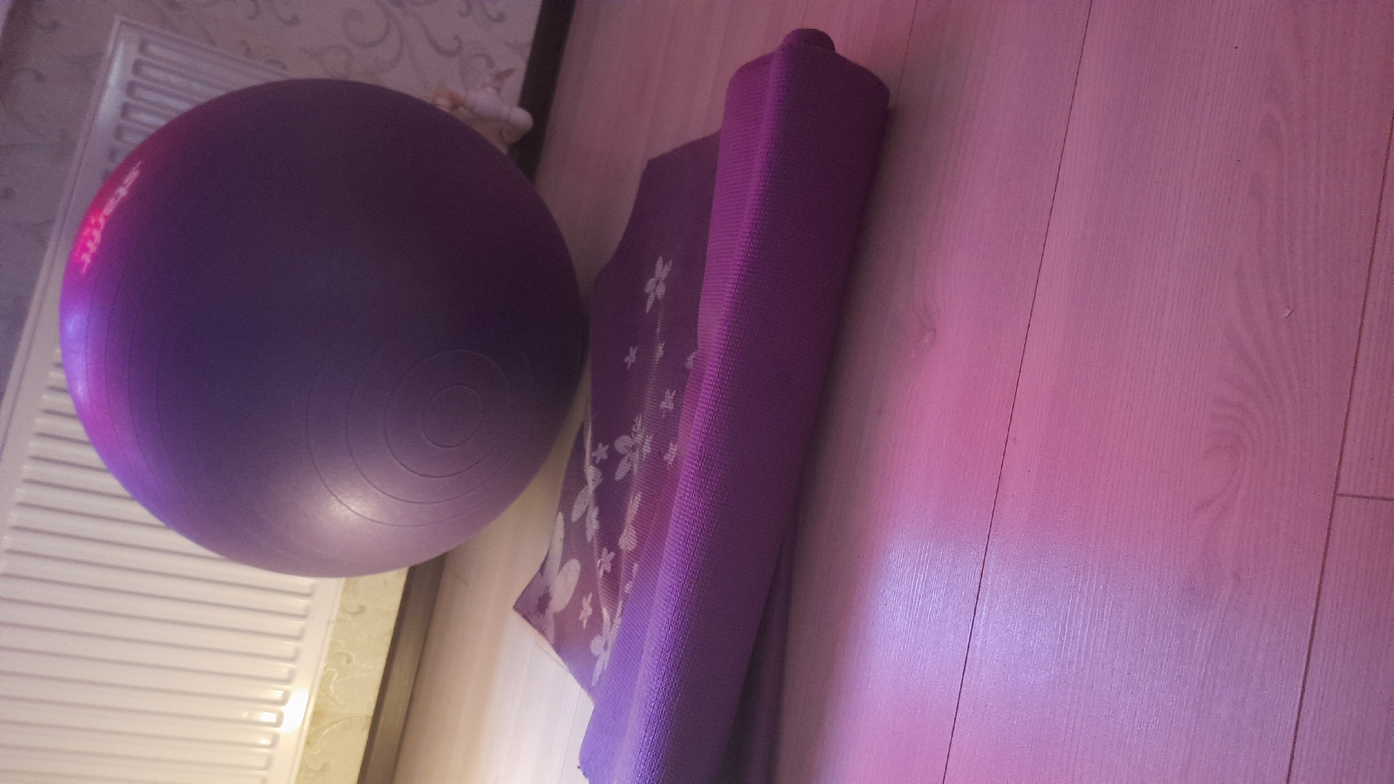 A purple exercise ball rests on a wooden floor next to a rolled-up purple yoga mat adorned with white floral patterns. The yoga mat is positioned in the foreground, while the exercise ball is in the middle ground. In the background, a white radiator and a patterned wallpaper are visible. The scene is illuminated by a pinkish light, creating a warm and inviting atmosphere. The image appears to be taken indoors, likely in a residential setting. The specific location in Desnogorsk, Russia cannot be determined from the visual information alone.