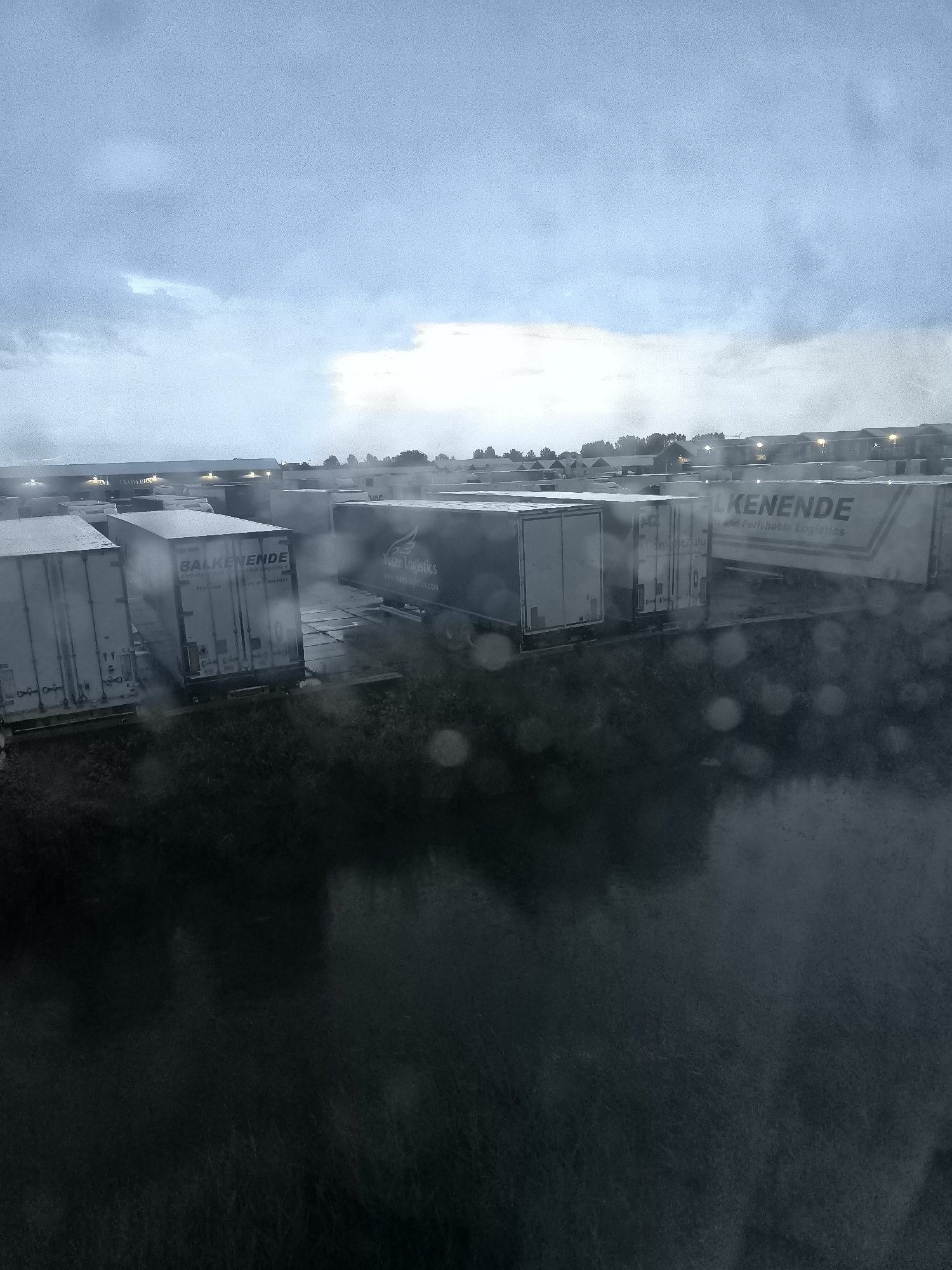 The image shows a view of a logistics company's yard in Uithoorn, Netherlands, on a cloudy and rainy day. Several white refrigerated trailers are parked in rows. One trailer has "BALKENENDE" and "Fresh & Perishable Logistics" printed on its side. Another trailer displays "Yusen Logistics" with a logo. In the background, buildings with lit windows are visible, suggesting a business complex. The foreground is dominated by a dark, wet surface, possibly a road or pavement, with numerous water droplets, indicating rain. The sky is overcast with diffused light.
