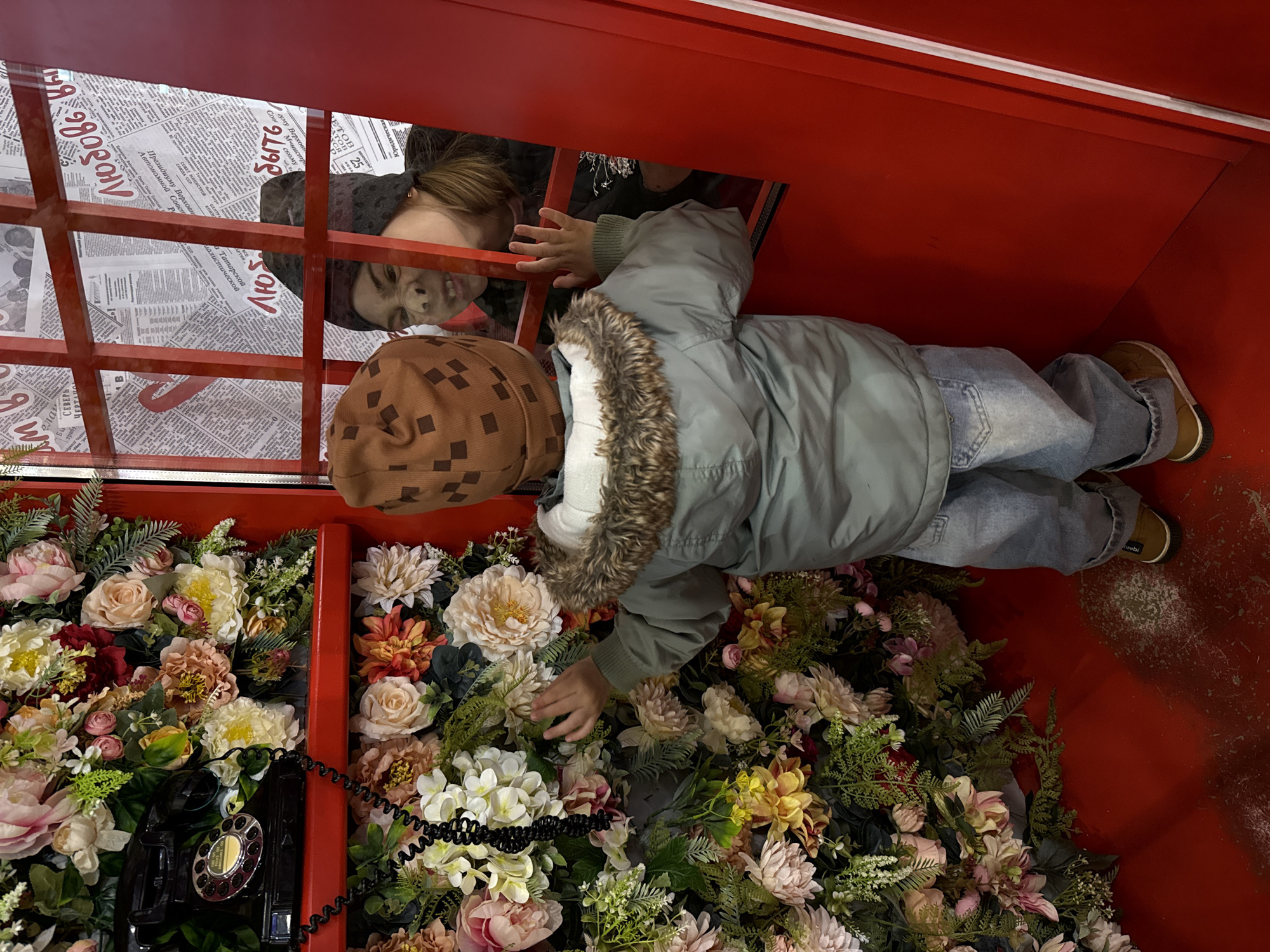 The image displays an artistic installation set within a red structure, reminiscent of a traditional phone booth. The space is filled with a dense arrangement of artificial flowers and green foliage in various shades of pink, cream, yellow, and red.
A child, wearing a light green hooded jacket with a faux fur trim, blue jeans, and brown boots, is positioned horizontally within the booth, reaching their hand into the mass of flowers. They also wear a brown cap with a checkered pattern. An antique-style black rotary telephone with a coiled cord is partially visible among the flowers in the lower-left corner.
Above the child, an adult's face is partially seen through a red grid or grate, looking down into the booth. The adult has a neutral expression and is wearing a dark head covering. Behind this grate, newspaper pages with Russian text are plastered, forming part of the background. Visible Russian words and phrases from the newspapers include "СОВЕТ МИНИСТРОВ ЦЕНТРА" (Council of Ministers of the Center), "Президиуму Верховн" (to the Presidium of the Supreme), "Татарской" (Tatar), and "Автономной Совет" (Autonomous Council). Numeric sequences like "2980" and "2L9S" are also present.
The scene conveys a playful and curious atmosphere, with the child exploring the vibrant floral display. The setting appears to be an indoor or protected environment, possibly in Korobovo, Russia, given the contextual information and visible Russian text.