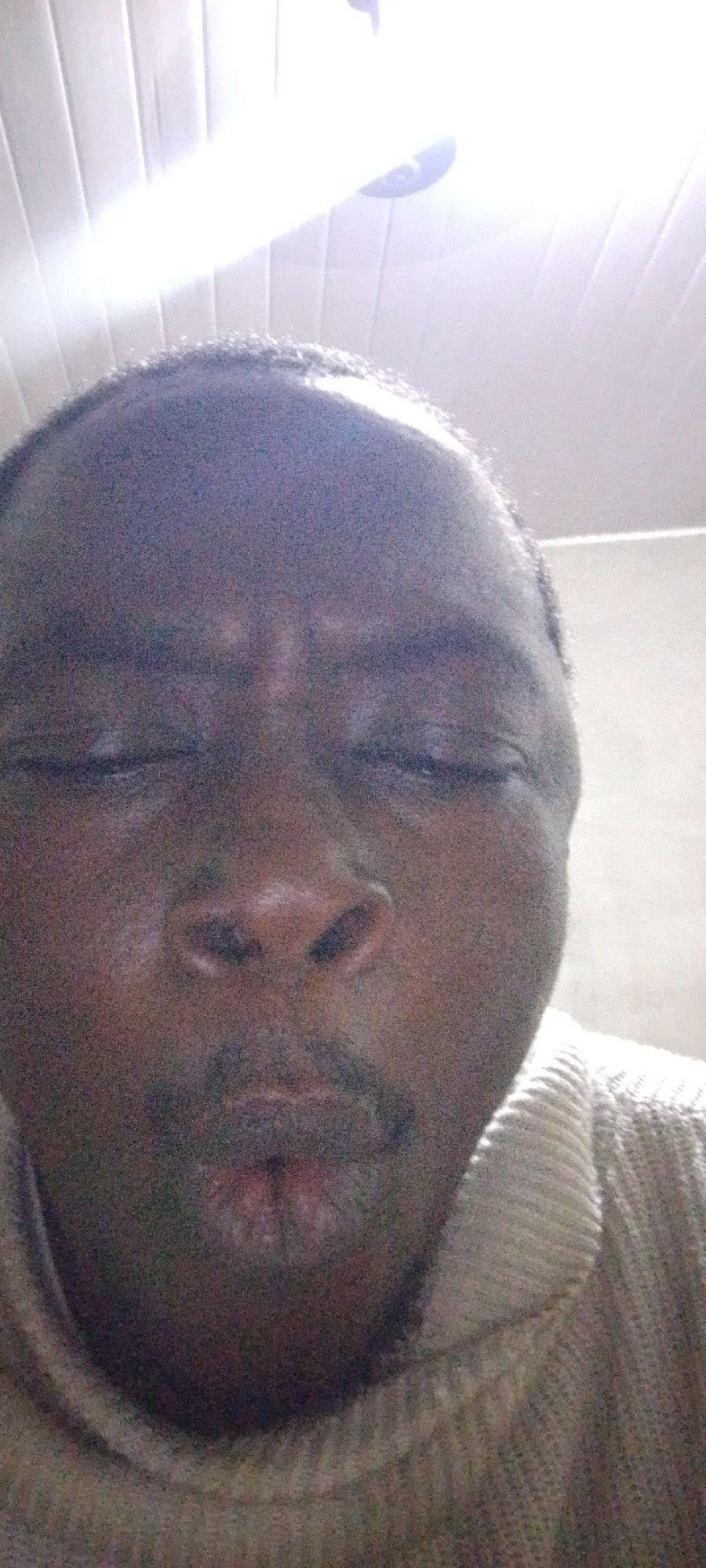 A close-up, poorly lit photograph features a person with dark skin, closed eyes, and a slight frown or pout. The person has a mustache and wears a light-colored sweater. The background is out of focus, with a light source or reflection visible at the top. There are no discernible details to indicate a specific location in Lokoja, Nigeria. The scene is focused entirely on the person's face.