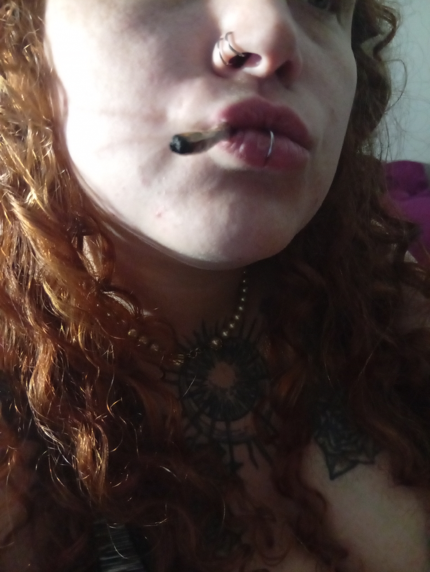 A close-up image shows a person with fair skin and curly, reddish-brown hair. They have a lip ring and a nose ring. A smoking joint is held in their mouth. Tattoos are visible on their chest and neck, along with a beaded necklace. The background is indistinct. The image cannot be identified as being in Buenos Aires.