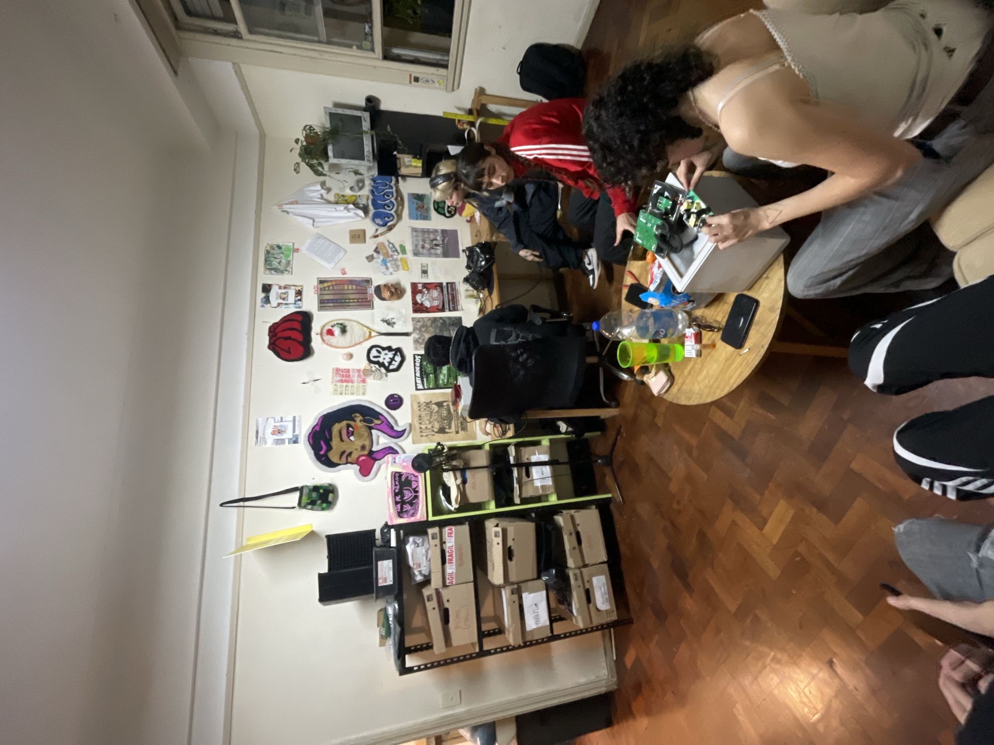 A low-angle indoor shot depicts a gathering of individuals in a cluttered room.
People: There are at least six individuals present in the room. One is wearing a white top and grey pants and working with a circuit board on a table. Another individual is wearing a red jacket with white stripes and is seated near another person in a black jacket. Another person is seated in a black chair facing away from the camera. Two other individuals are partially visible in the lower right corner of the frame.
Subjects: The subjects appear to be working or engaged in a project involving electronics, as evidenced by the circuit board and other electronic components on the table.
Objects: Notable objects include a round wooden table with various items on it, including bottles, keys, and a smartphone. There's also a black chair and a shelving unit stocked with cardboard boxes. The walls are adorned with various posters and stickers, and there is a window in the background.
Actions: The primary action is the manipulation of a circuit board, with other individuals observing or assisting. There is a clear interaction between the people working on the project.
Environment: The room has a lived-in appearance, with items scattered around. The floor is made of wooden planks. The room appears to be an apartment, judging by the decor and furniture.
Location: Based on the architectural style and room layout visible, it is reasonable to infer that the location is Buenos Aires, Argentina.