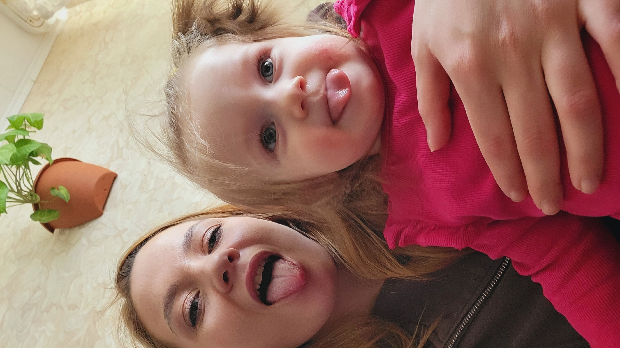 A woman and a toddler are shown from a low angle, their faces close together, both sticking out their tongues. The toddler is wearing a bright pink garment, and an adult hand is visible near the toddler. The woman has brown hair and is wearing a dark-colored jacket with a zipper. A potted plant is present on the left side of the frame. The setting appears to be indoors, with patterned wallpaper or flooring visible. The location is likely Chaikovsky, Russia, based on the prompt.