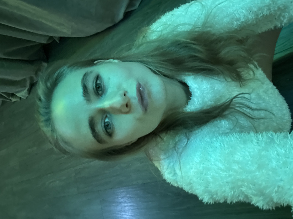 A fair-skinned woman with light hair and blue eyes gazes directly at the camera. She wears a fluffy, white sweater. The lighting has a greenish-blue cast, altering skin tones. A portion of what appears to be a brown velvet cushion is visible in the upper left. The background appears to be a dark wood floor. The image is rotated slightly.