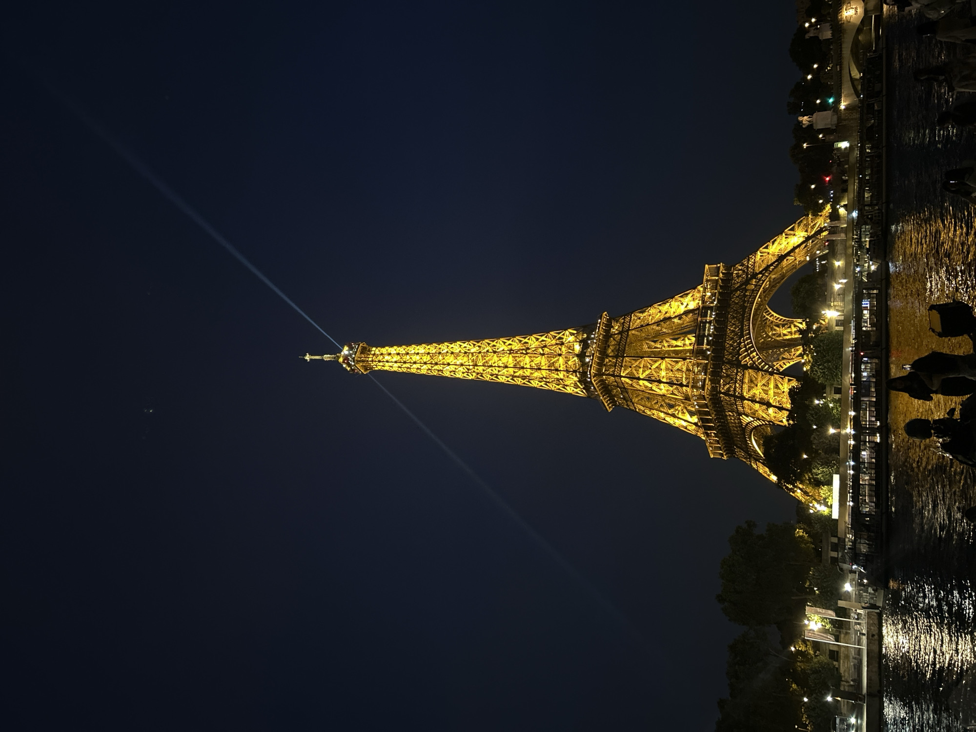 This is a nighttime, vertical, full shot of the Eiffel Tower, illuminated in warm yellow light, in Paris, France. The iconic iron lattice structure dominates the frame, with a searchlight beam extending upwards into the dark blue sky from its summit. Below the tower, the Seine River reflects the golden lights of the tower and city. Several boats are docked or cruising on the river. Along the riverbank, trees and streetlights are visible, as are silhouetted figures of people. The overall impression is one of a classic Parisian nighttime scene.