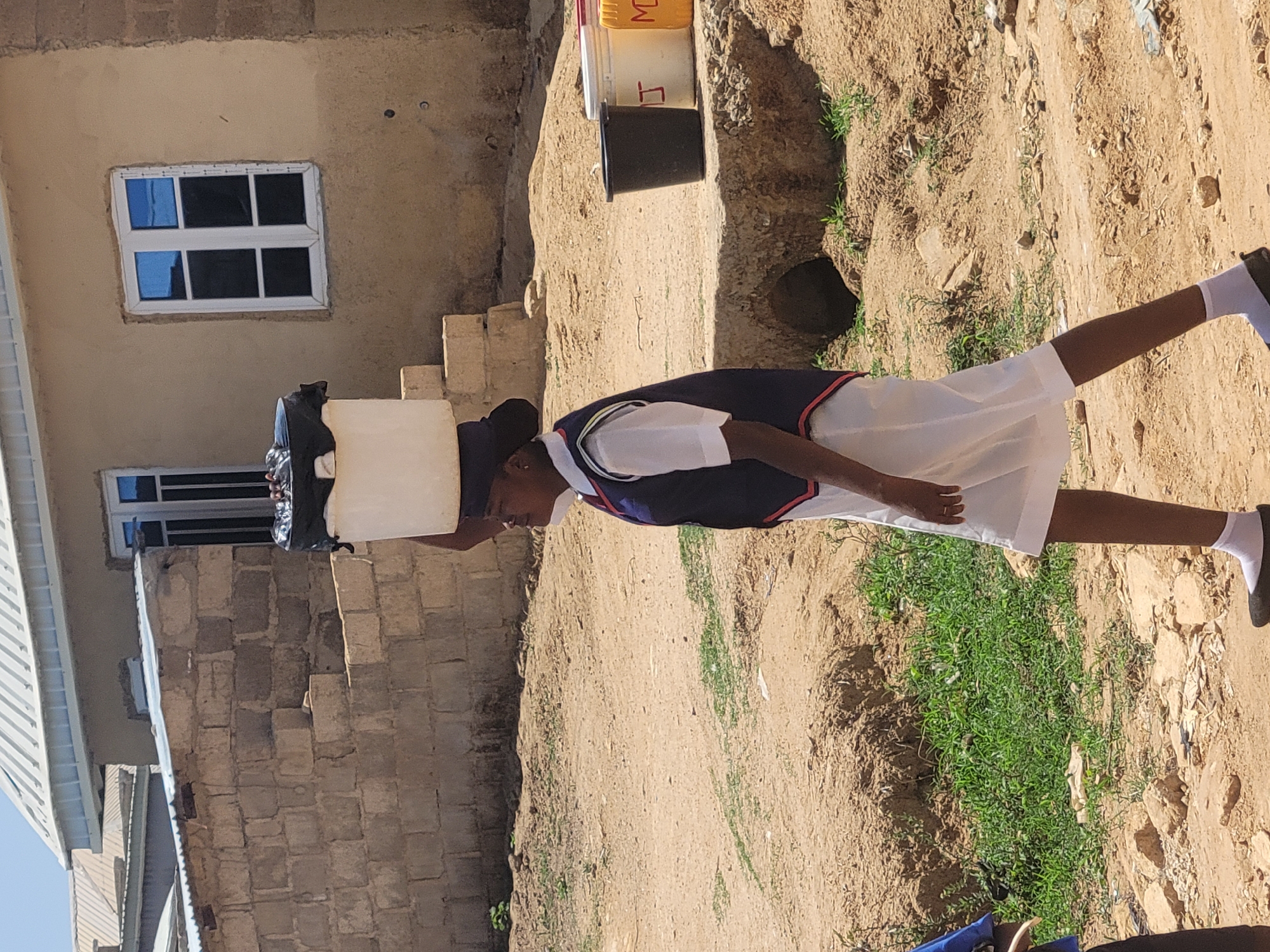 A person in a white school uniform, wearing white socks and black shoes, is carrying a white container covered with black plastic on their head. The person is walking on a dirt path with some grass and rocks on the side. Behind the person is a building with a window. There are some buckets stacked near the building. The location is likely in Jos, Nigeria, based on the attire and surroundings.