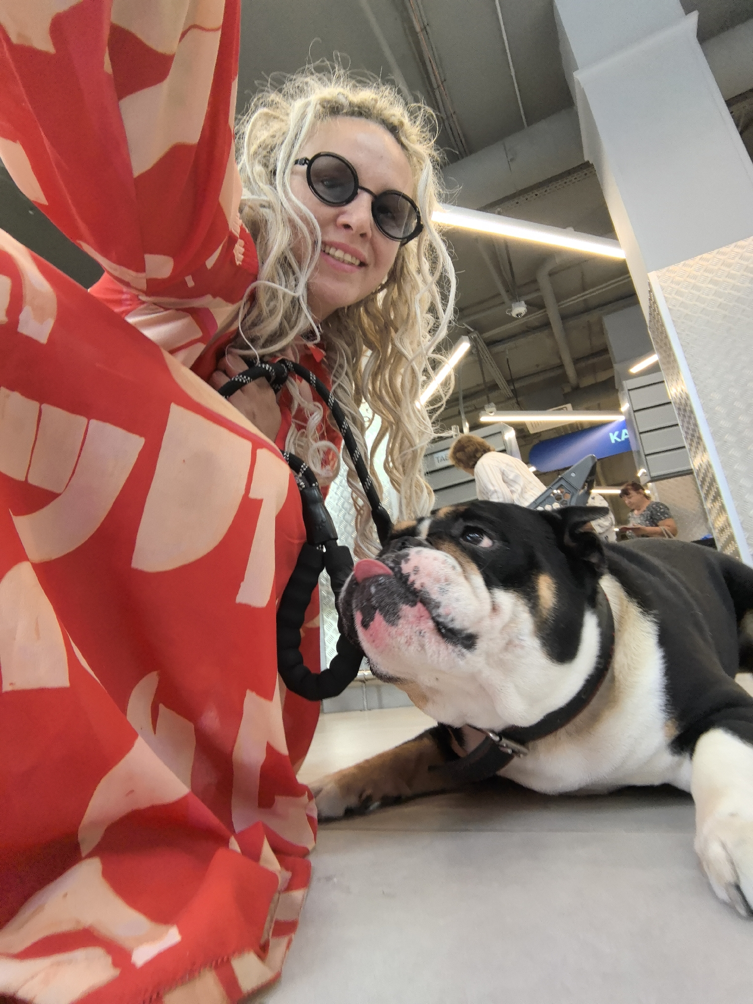 The image shows a woman with blonde curly hair wearing round sunglasses and a red dress with a white pattern. She is holding the leash of a black, white, and tan bulldog. The dog is lying on the floor and licking its lips, looking up at the woman. In the background, there are other people and an indoor environment that appears to be a public space, possibly a shopping mall or a train station, with metal structures and lighting fixtures visible on the ceiling. There is a sign with the Cyrillic letters "КА" on it. While the specific location in Nizhny Novgorod, Russia, is not definitively identifiable from the visual cues alone, the overall context suggests an urban public indoor setting.