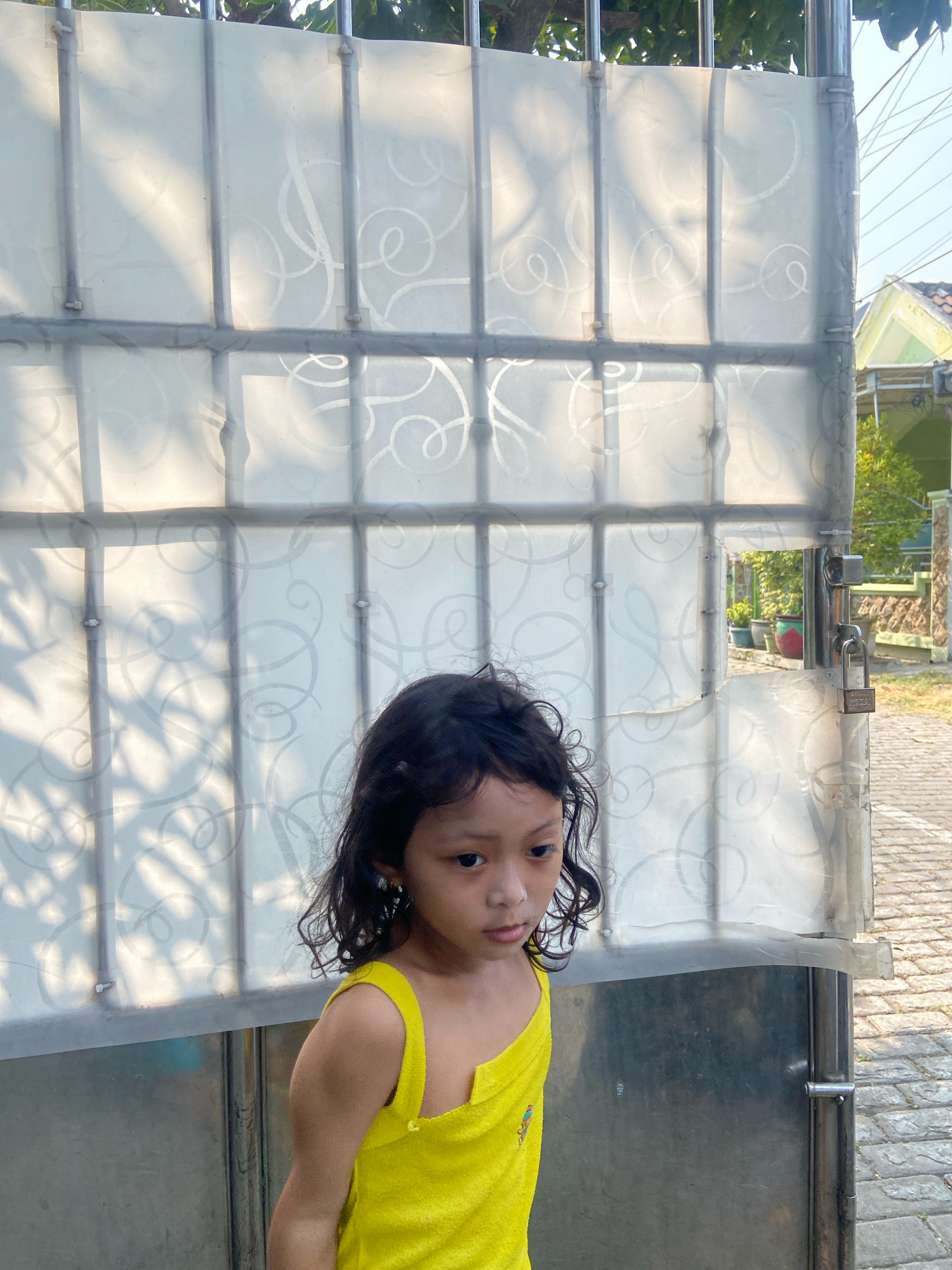 A young girl with dark, curly hair stands in front of a translucent gate in Gelam, Indonesia. She is wearing a bright yellow one-piece garment. The gate is made of metal bars and translucent panels with a swirling pattern. Sunlight casts shadows on the gate, creating a dappled effect. Behind the gate, a paved walkway and some greenery are visible. The overall scene appears to be outdoors in a residential area.