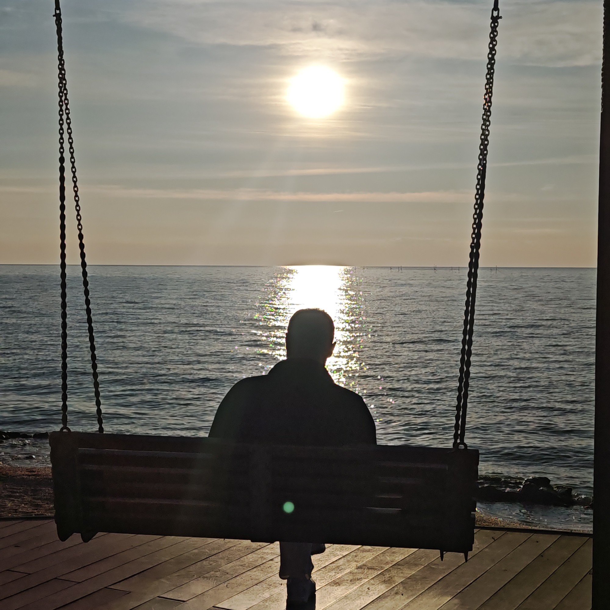 The image depicts a person sitting on a swing facing the sea at sunset. The location is likely near or in городское поселение Щелкино, Ukraine, given the prompt parameters. The person is silhouetted against the bright sun reflecting off the water. The swing hangs from chains attached to an unseen structure. The ground is a wooden deck. The sea is calm with slight ripples, and the sky is clear with a bright sun.