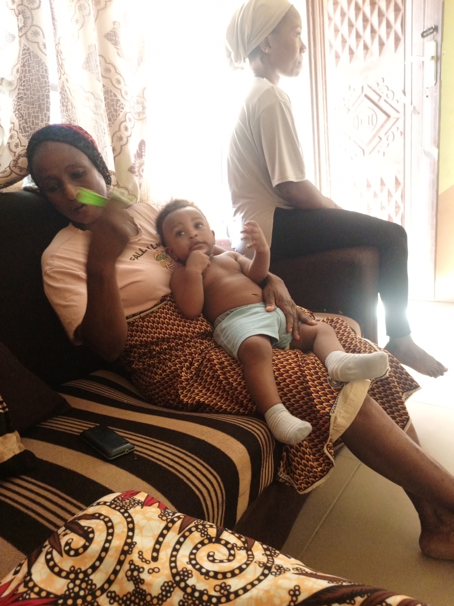 A woman sits on a striped couch in Unknown, Unknown, holding a baby on her lap. The baby is wearing a light blue diaper and white socks. The woman is wearing a pink t-shirt, a patterned skirt, and a head covering. She is holding a green object in her hand. Another person, also wearing a head covering, sits in the background near a door. A wallet lies on the couch beside the woman. Curtains are visible in the background.