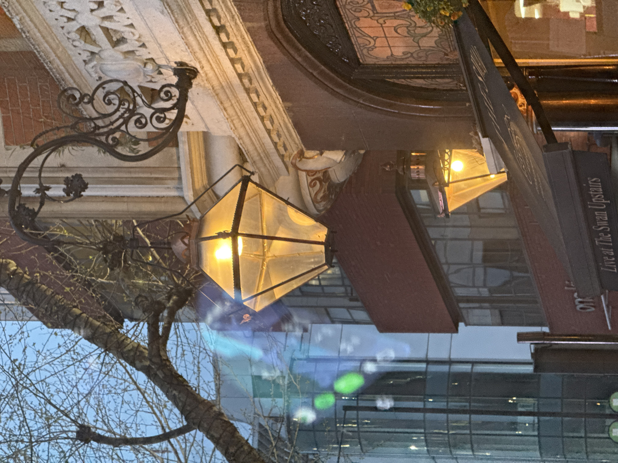 The image depicts a street scene in London, United Kingdom. The focus is on an ornate street lamp attached to a building facade with intricate architectural details. A sign reading "Live at the Swan Upstairs" is visible to the right. A tree with bare branches is in the foreground, partially obscuring the view of the building beyond, which appears to be a modern structure with large windows. The sky is pale blue, and the lighting suggests it is either dusk or dawn. The overall impression is of a juxtaposition between historic and modern architecture in an urban setting.