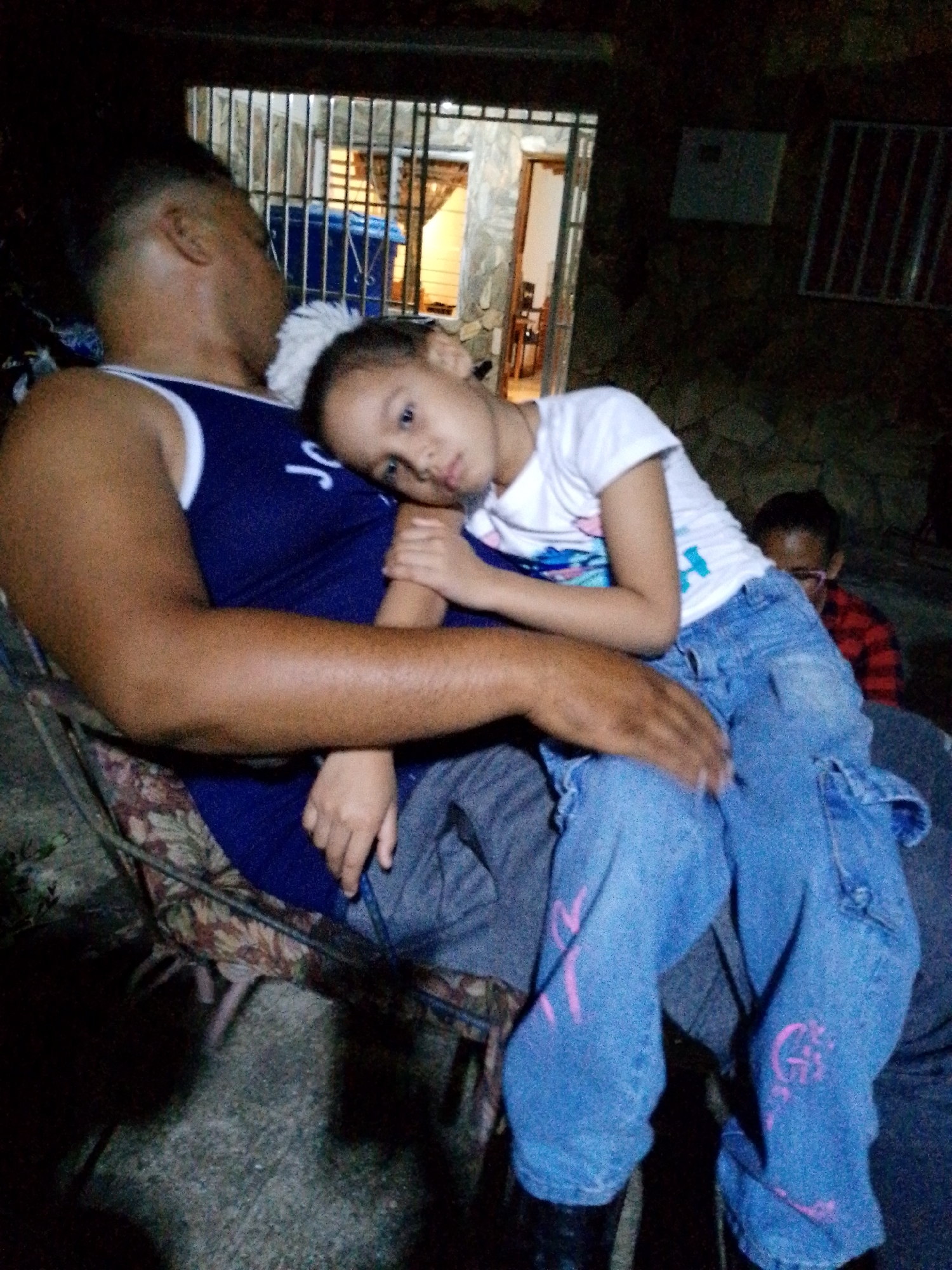 The image depicts a close-up view of a man holding a young child, possibly his daughter, on his lap. They are seated outdoors at night, with a dimly lit background suggesting a residential area in Juan Griego, Venezuela. The man is wearing a dark blue tank top and jeans, while the child is dressed in a white t-shirt and blue jeans with pink markings. The child's head is resting on the man's shoulder, indicating a moment of rest or comfort. Another person, a young boy wearing glasses and a red and black plaid shirt, is visible in the background, adding to the familial atmosphere. The foreground shows a portion of a patterned chair and the rough texture of concrete ground, hinting at an outdoor seating arrangement.